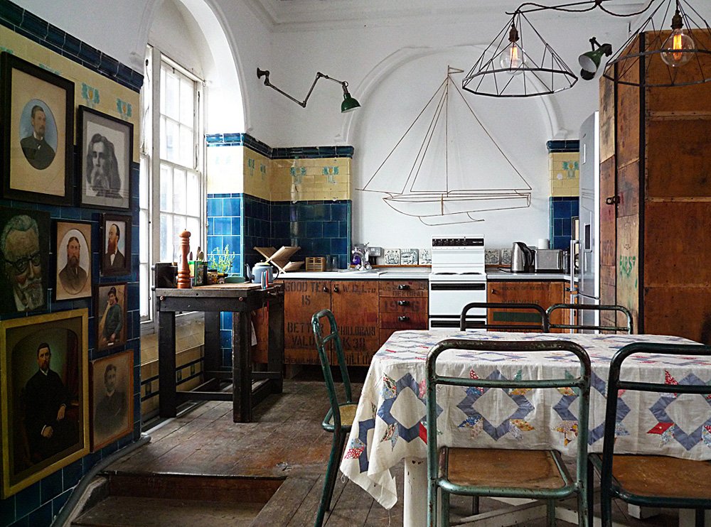A cozy kitchen with a table covered in a patterned tablecloth, surrounded by mismatched chairs. The wall features portraits and a wire outline of a sailboat. There are kitchen appliances and utensils on the counter, with a window letting in natural l