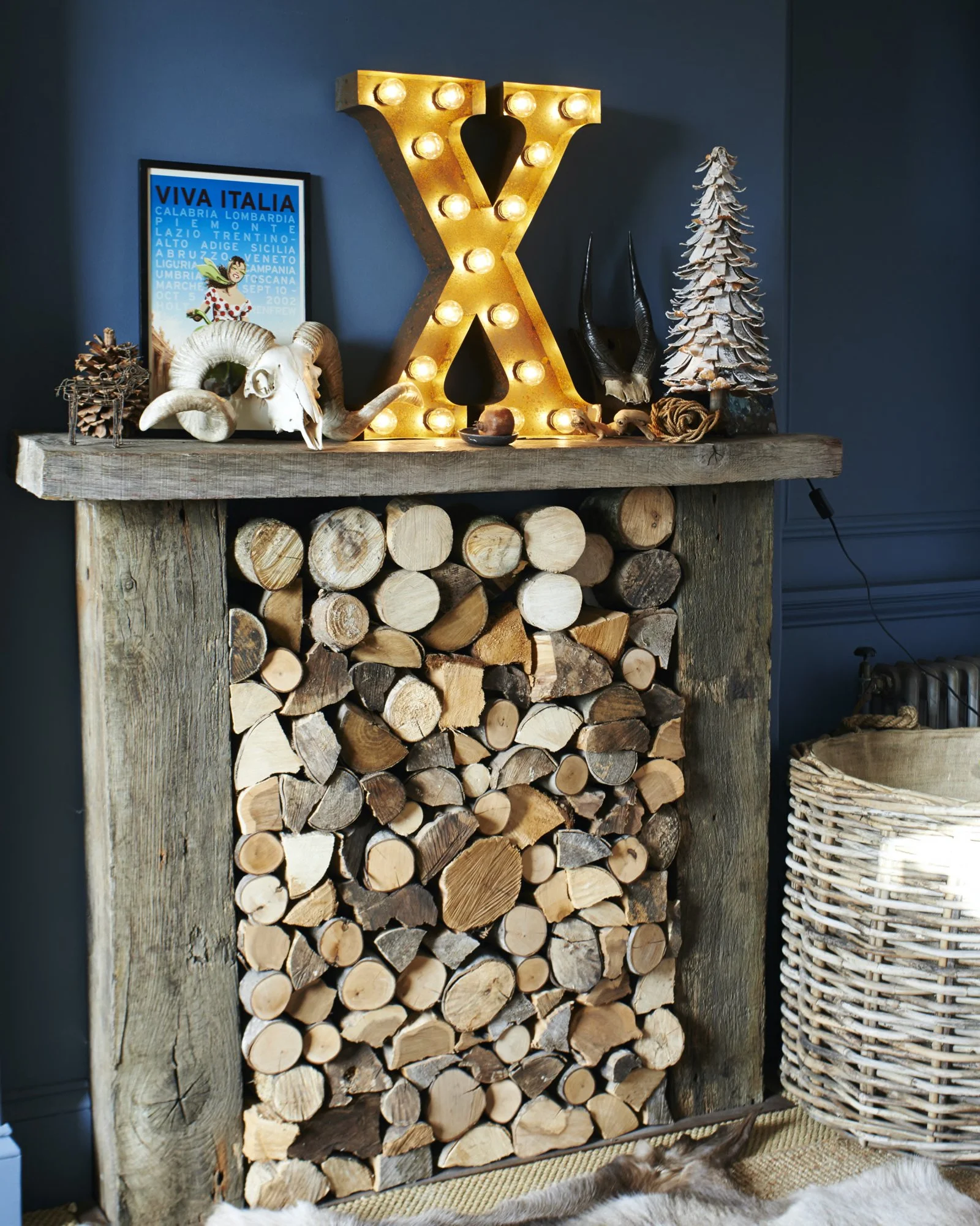Decorative shelf with logs, a large illuminated letter 'X', and various ornaments including animal skulls, a small pine tree, and a framed poster, all against a blue wall.