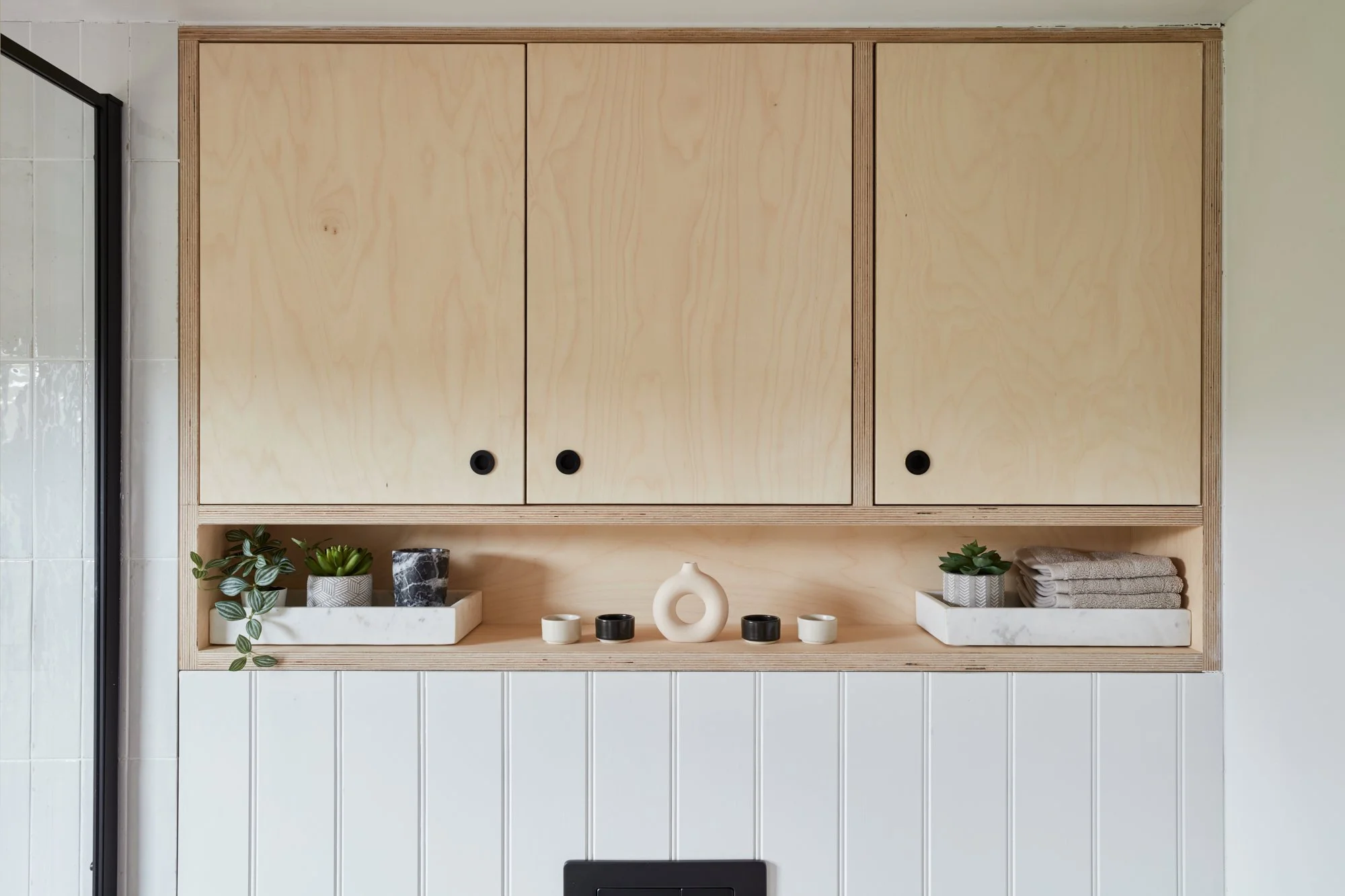 Wooden cabinet with closed doors, decorative plants, bowls, and towels on a shelf underneath, and a tiled wall in the background.