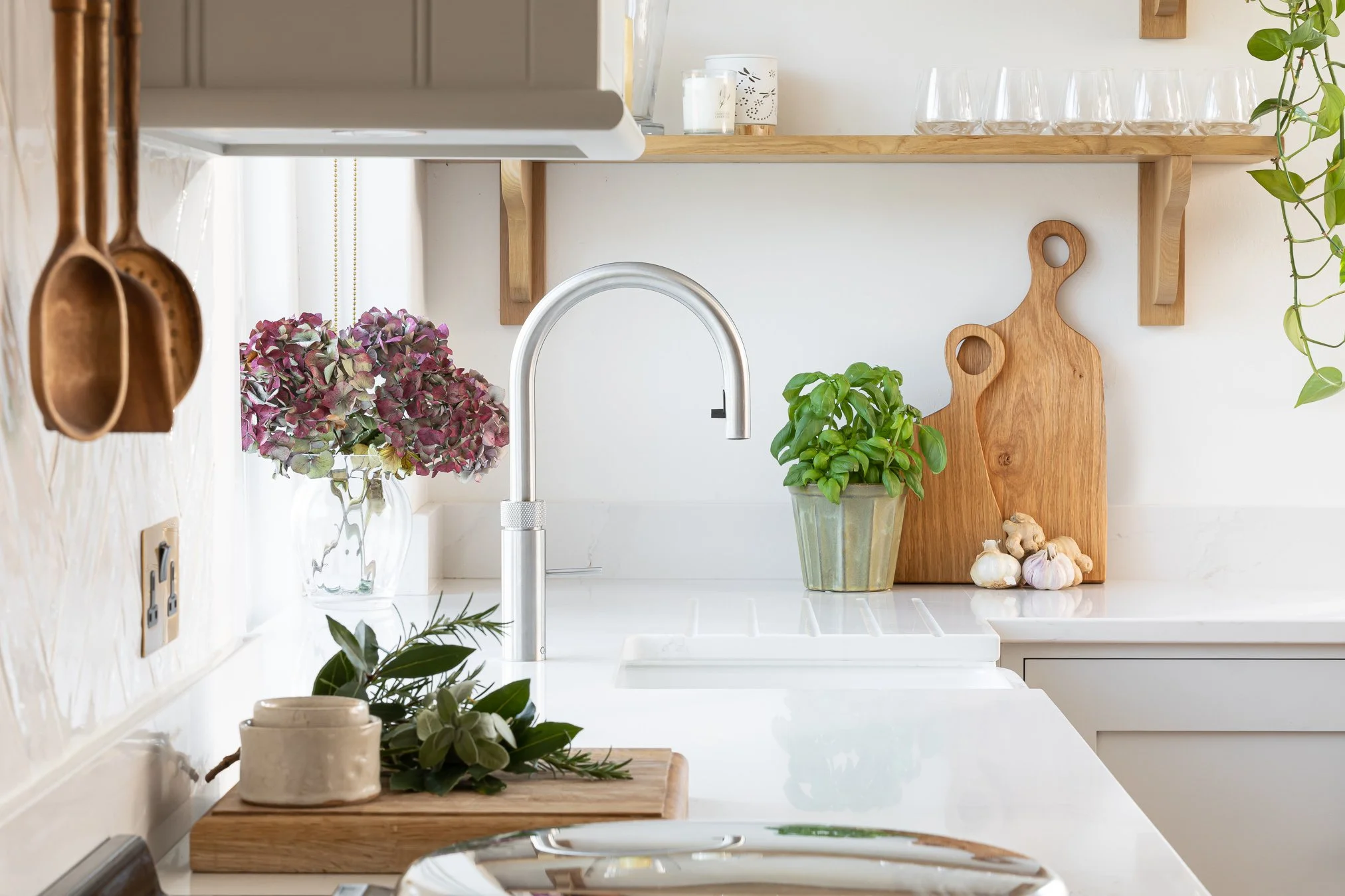 A clean, modern kitchen with a white countertop, a silver faucet, and wooden shelves. There are potted plants, garlic, and decorative items on the counter, with hanging copper utensils and glassware on the shelves.