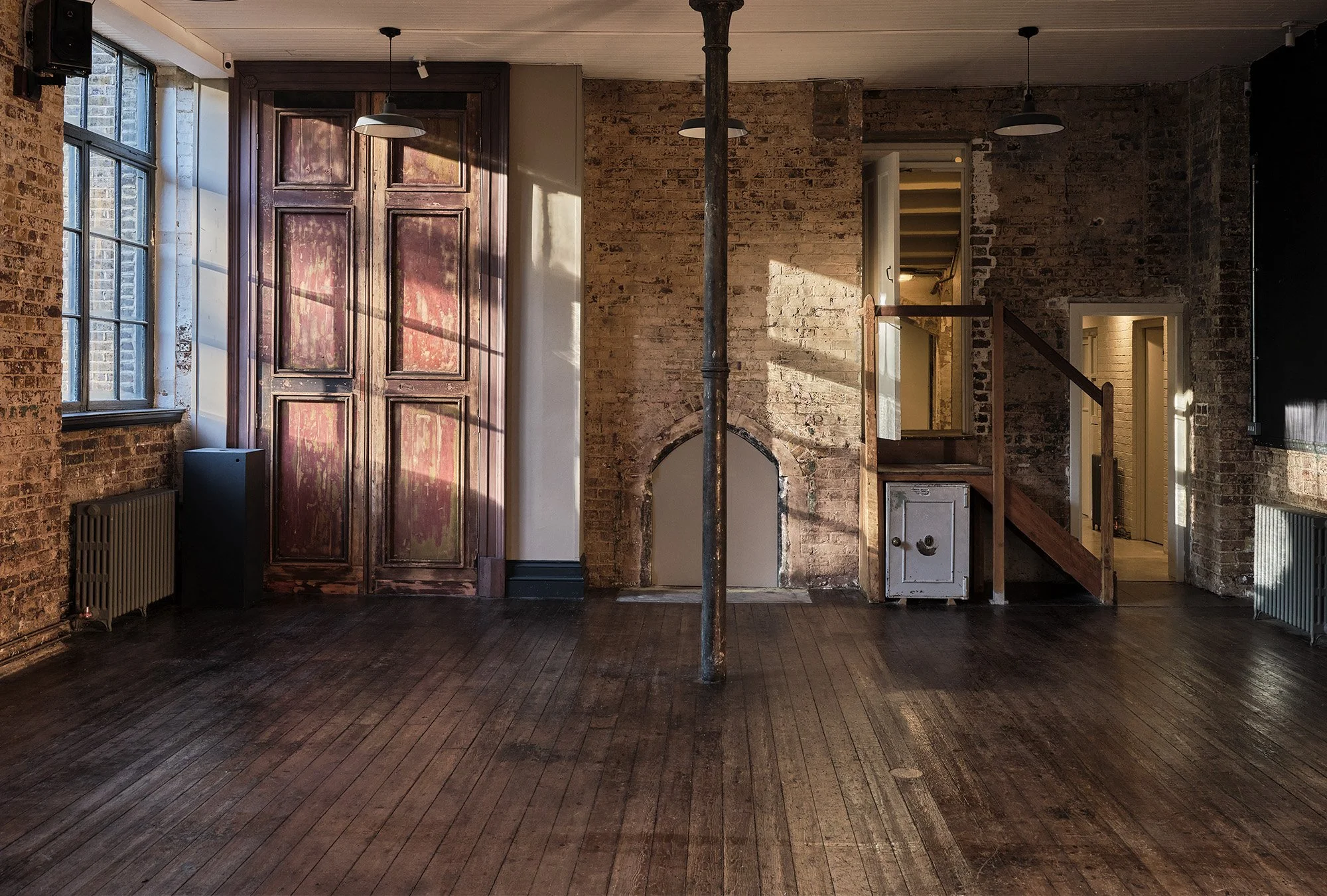 Empty room with brick walls, large windows, wooden floors, a metal pole, and an industrial chimney. Stairs lead to an upper level, and sunlight creates shadows on the walls.