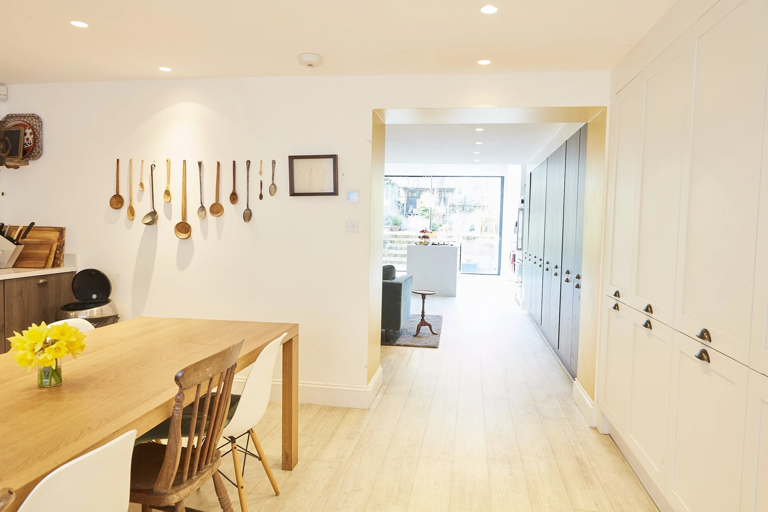 Bright dining area with a wooden table, white and wooden chairs, a vase of yellow flowers, kitchen cabinets with wooden knobs, and a wall decorated with hanging wooden spoons