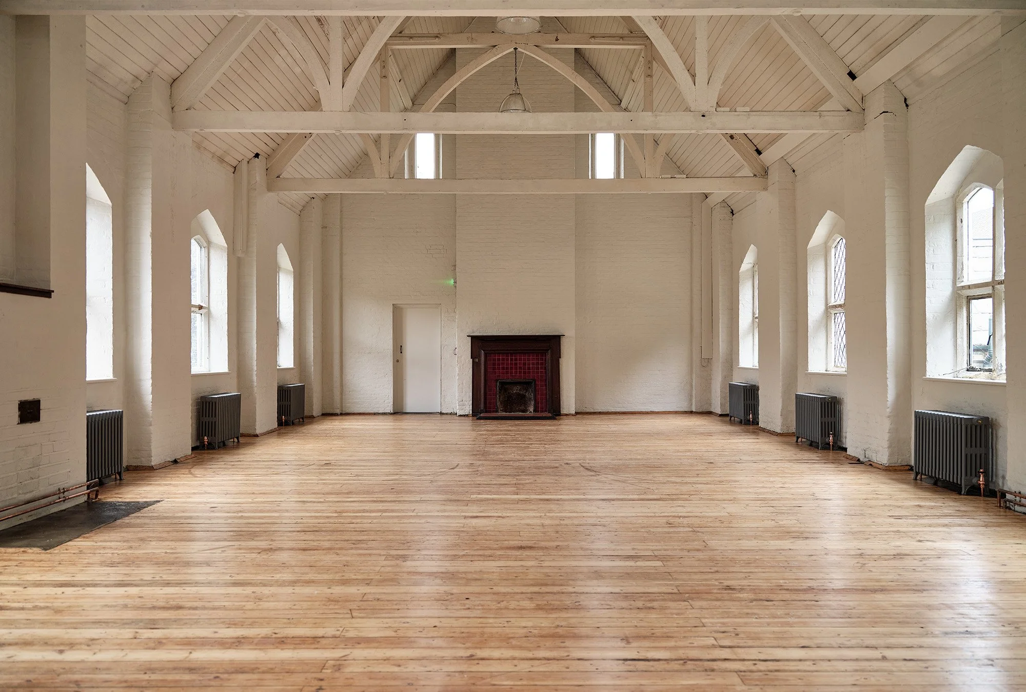 Large, empty room with white painted brick walls, arched wooden ceiling, wooden floor, and several windows with black radiators underneath. There is a fireplace with red brick tiles in the center of the back wall and a white door to the left of it.