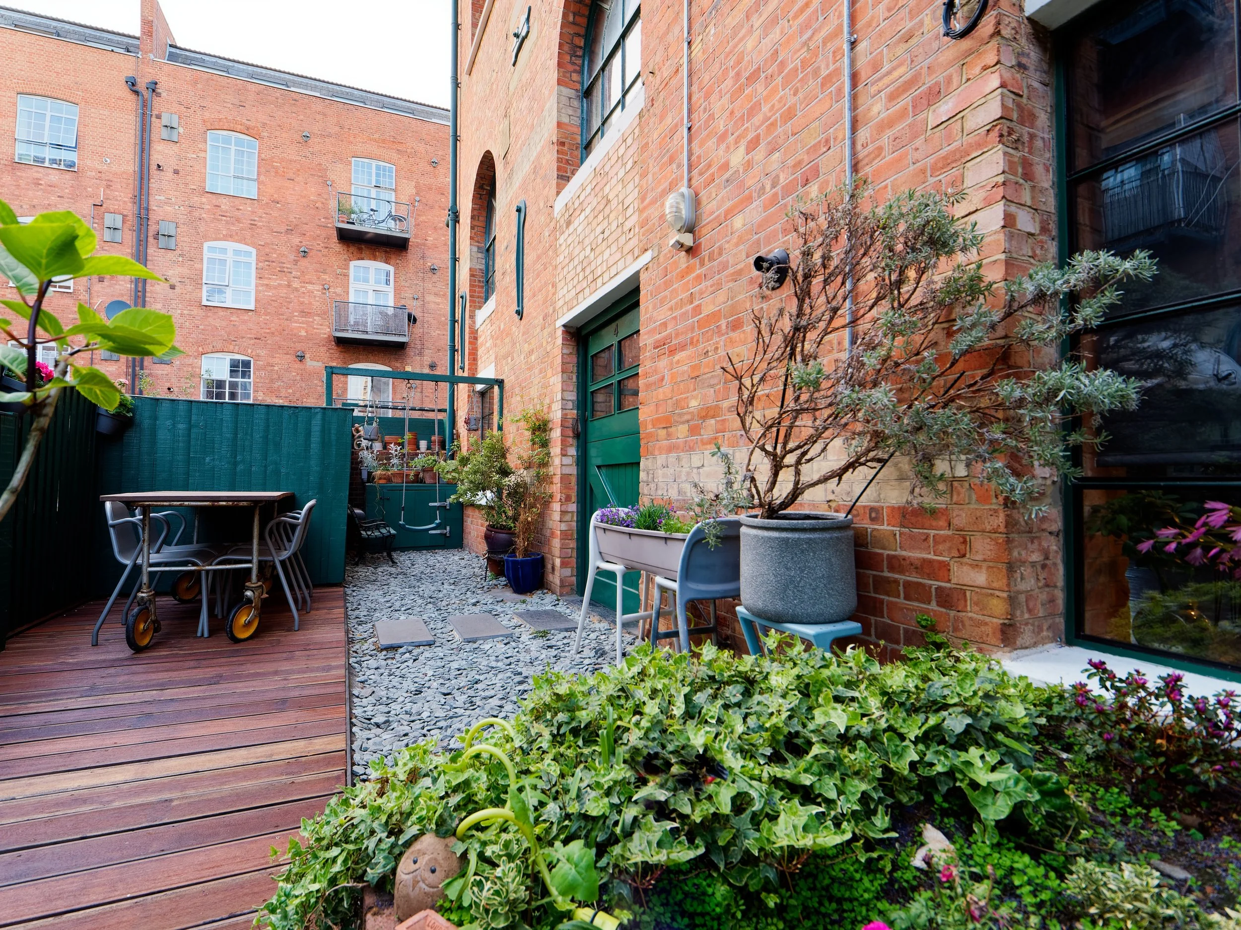 View of a small urban backyard with a wooden deck, potted plants, a table with chairs, and a brick building wall with large windows.
