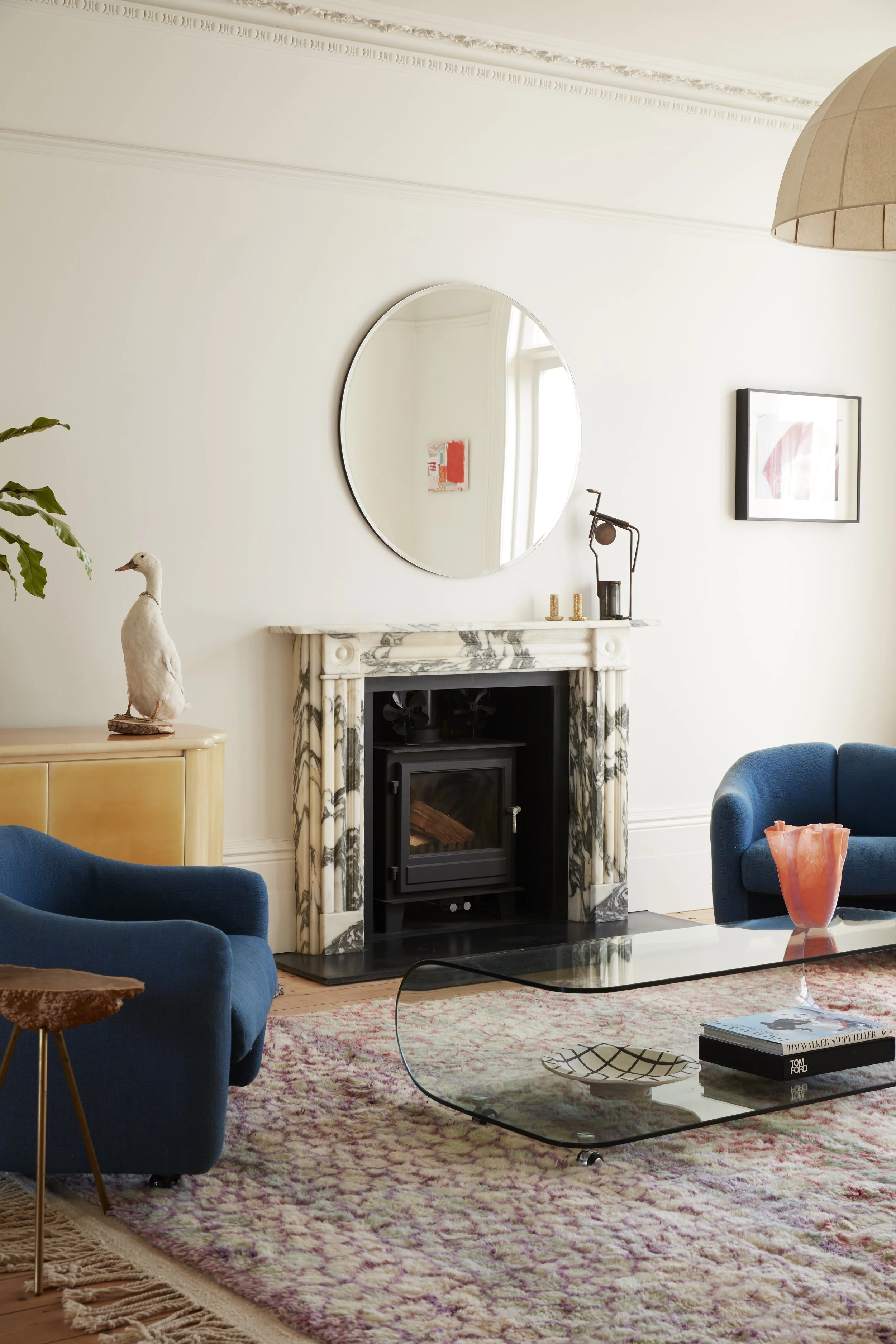 A cozy living room with a marble fireplace, a round mirror on the wall, a fireplace with a wood stove, a blue armchair, a glass coffee table, a pink vase, and various art pieces on the wall.