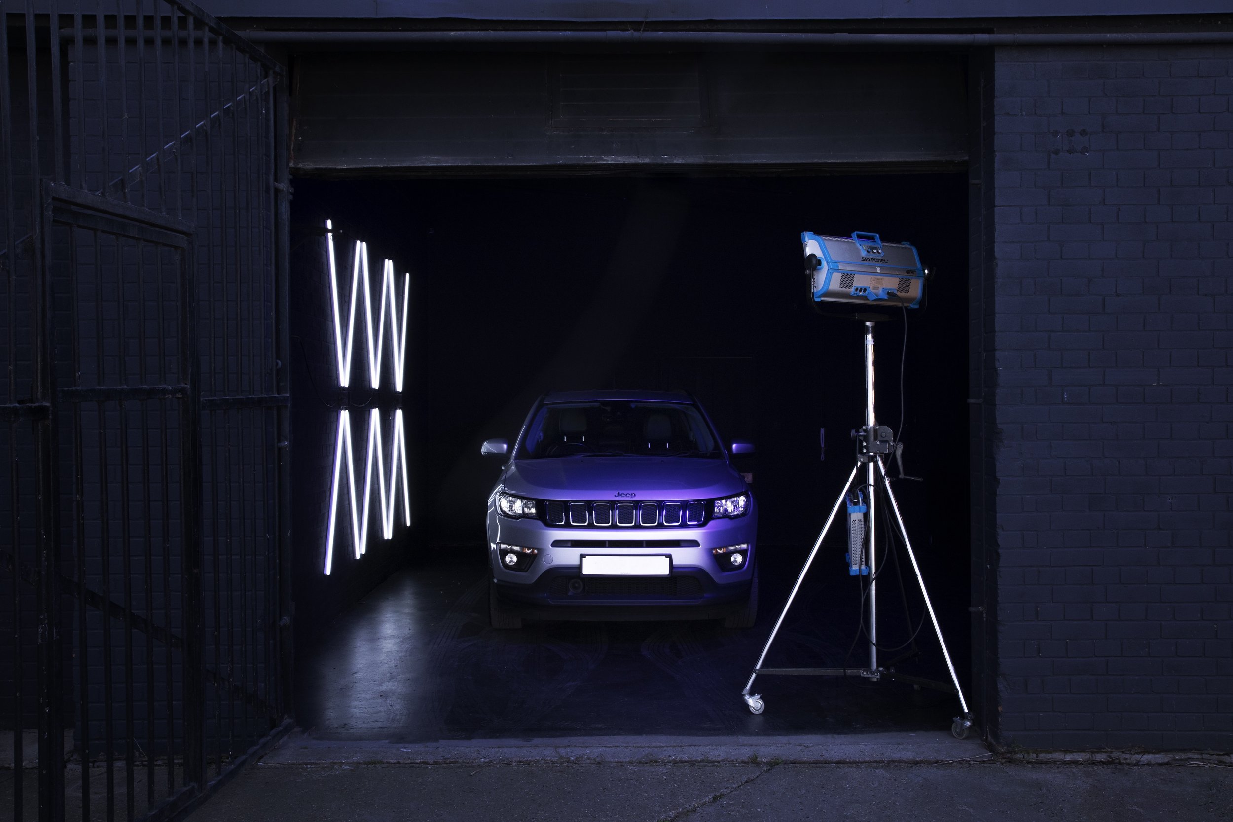 A silver Jeep SUV inside a dark garage with black brick walls, illuminated by a large softbox light on a stand to the right, and purple neon lights on the left wall.
