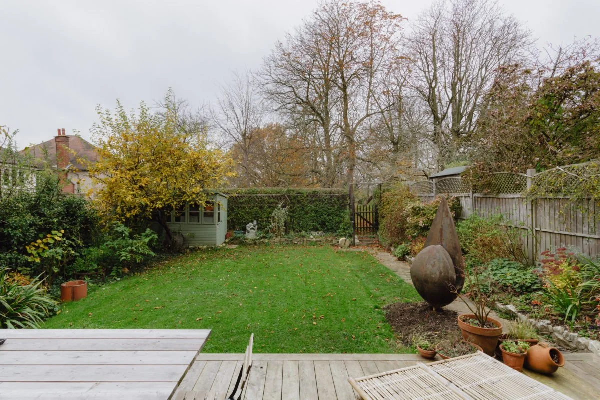 A backyard garden with a lawn, trees, potted plants, and decorative sculptures, surrounded by a wooden fence and privacy hedges in autumn.