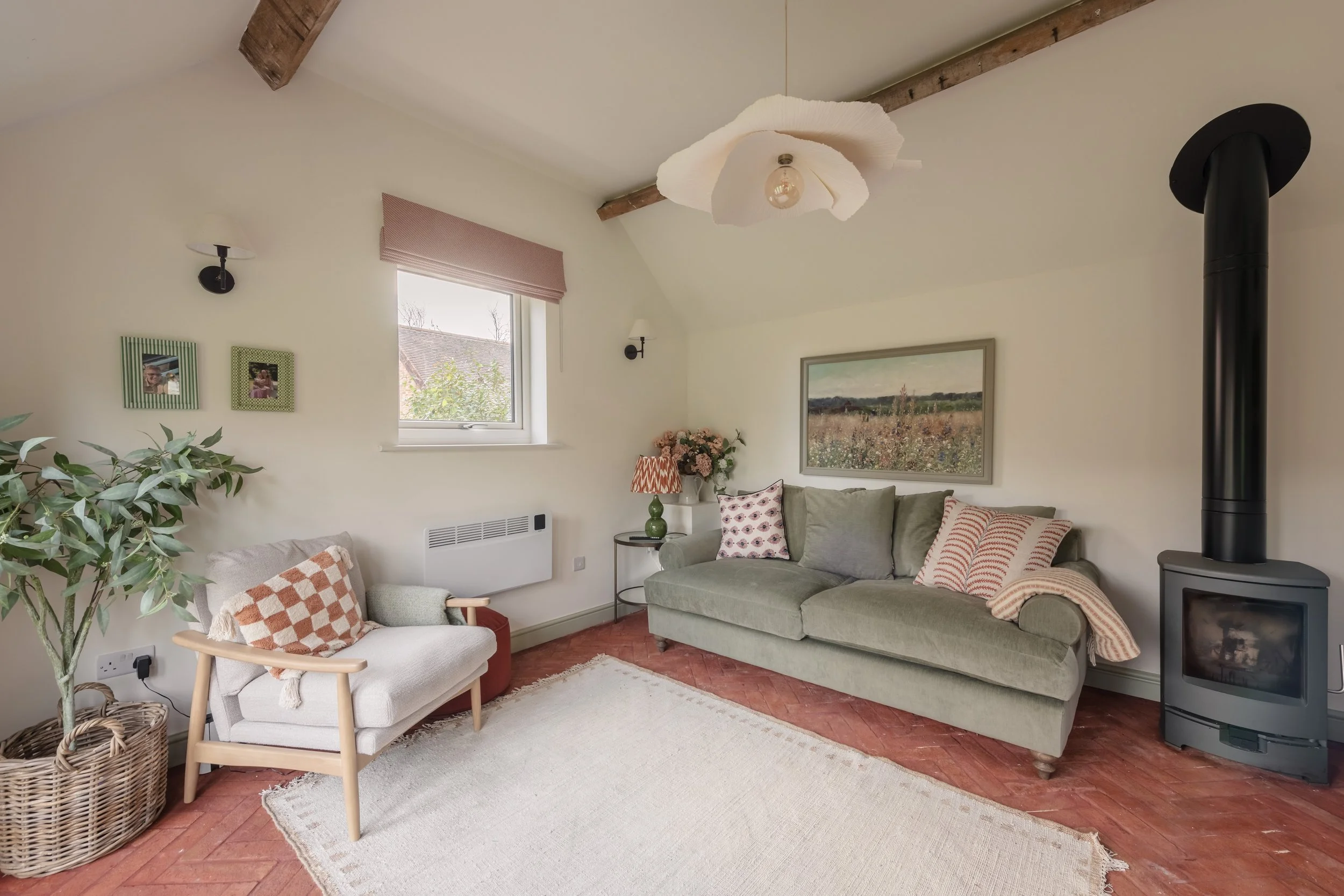 Living room with a green sofa, a cream armchair, a wooden armchair with checkered pillow, a small side table, a vase of flowers, framed wall art, a window with pink shade, a beige rug, a wood stove, and exposed wooden ceiling beams.