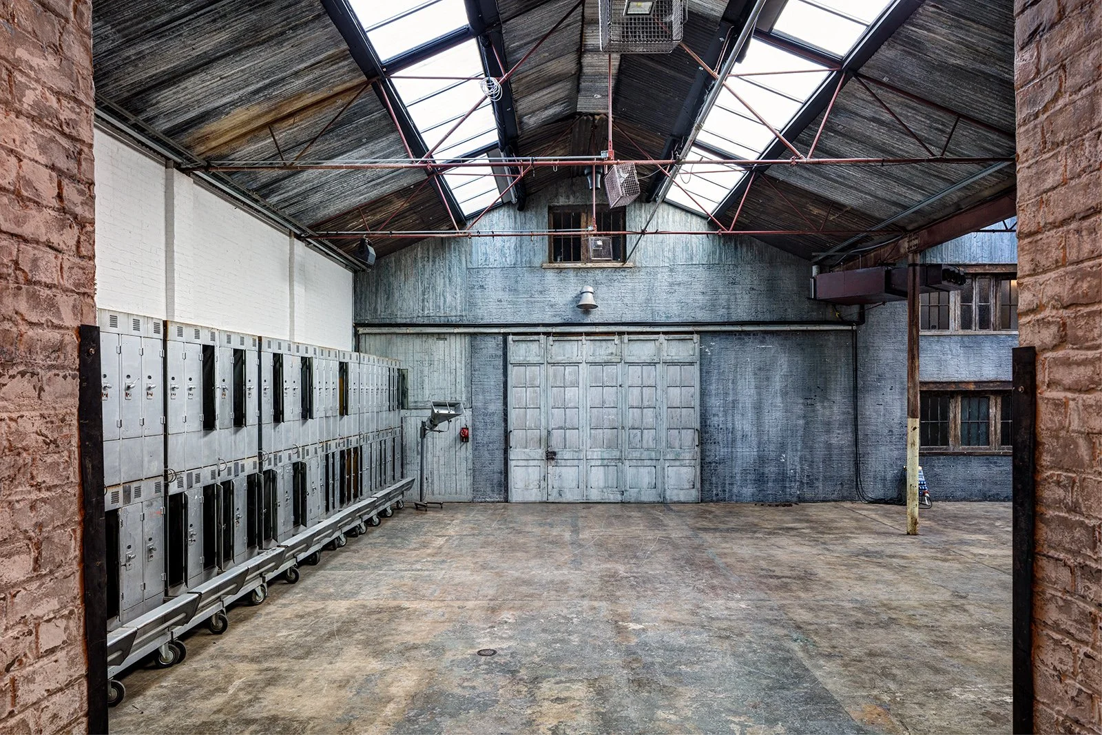 Empty industrial warehouse with metal lockers along the left side and a large door in the back wall, high skylight windows, and exposed brick walls.