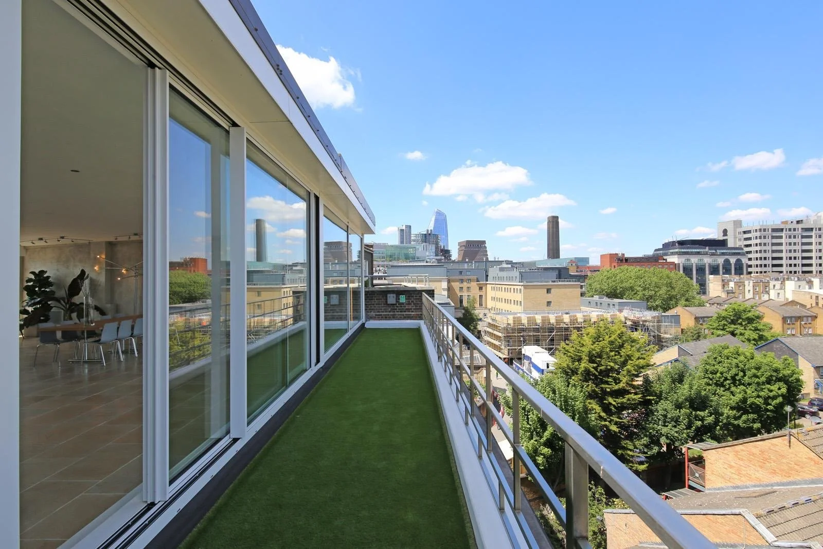Balcony with artificial grass flooring overlooking a city skyline with tall buildings and scattered trees under a blue sky.