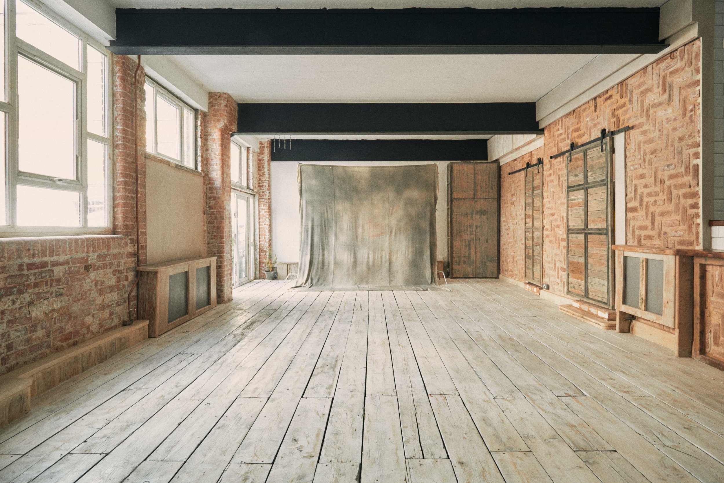An empty room with large windows, exposed brick walls, and wooden floors, featuring a backdrop or curtain in the middle and sliding barn doors on the right wall.