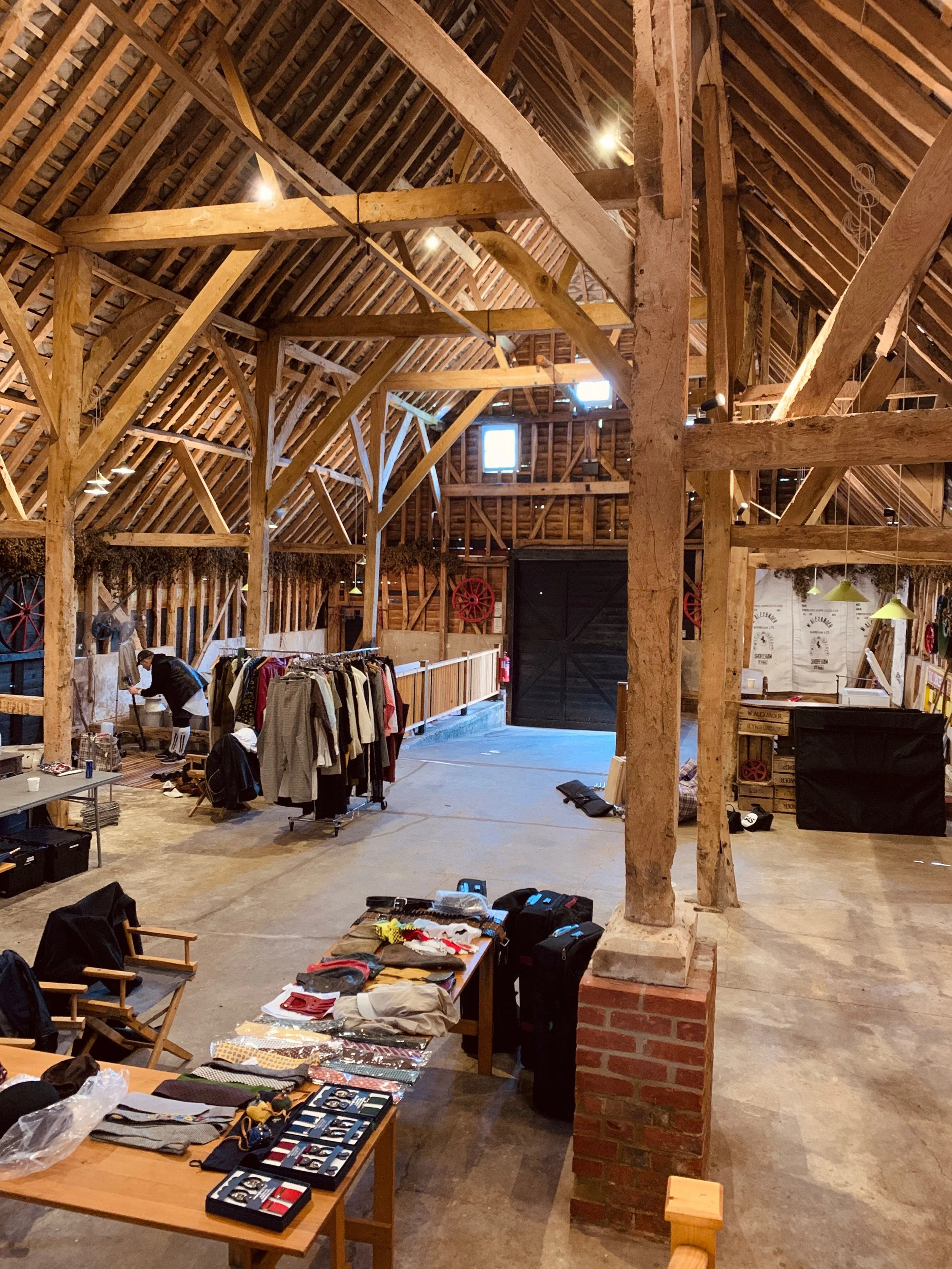 Interior view of a rustic, large barn converted into a marketplace or event space with exposed wooden beams, brick pillars, and display tables filled with clothes, accessories, and other items. A person is seen browsing near clothing racks, and vario