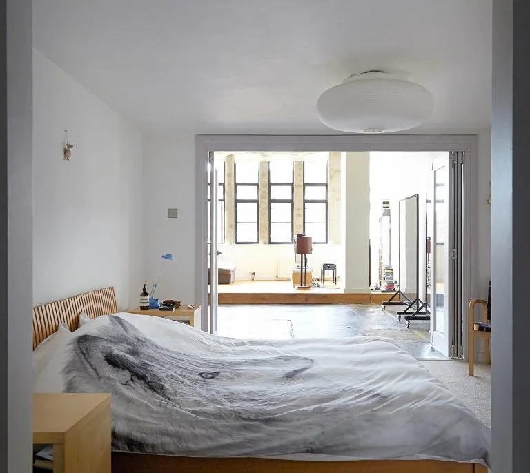 Bedroom with a bed, side table, and a lamp. The room leads to a bright living space with large windows.