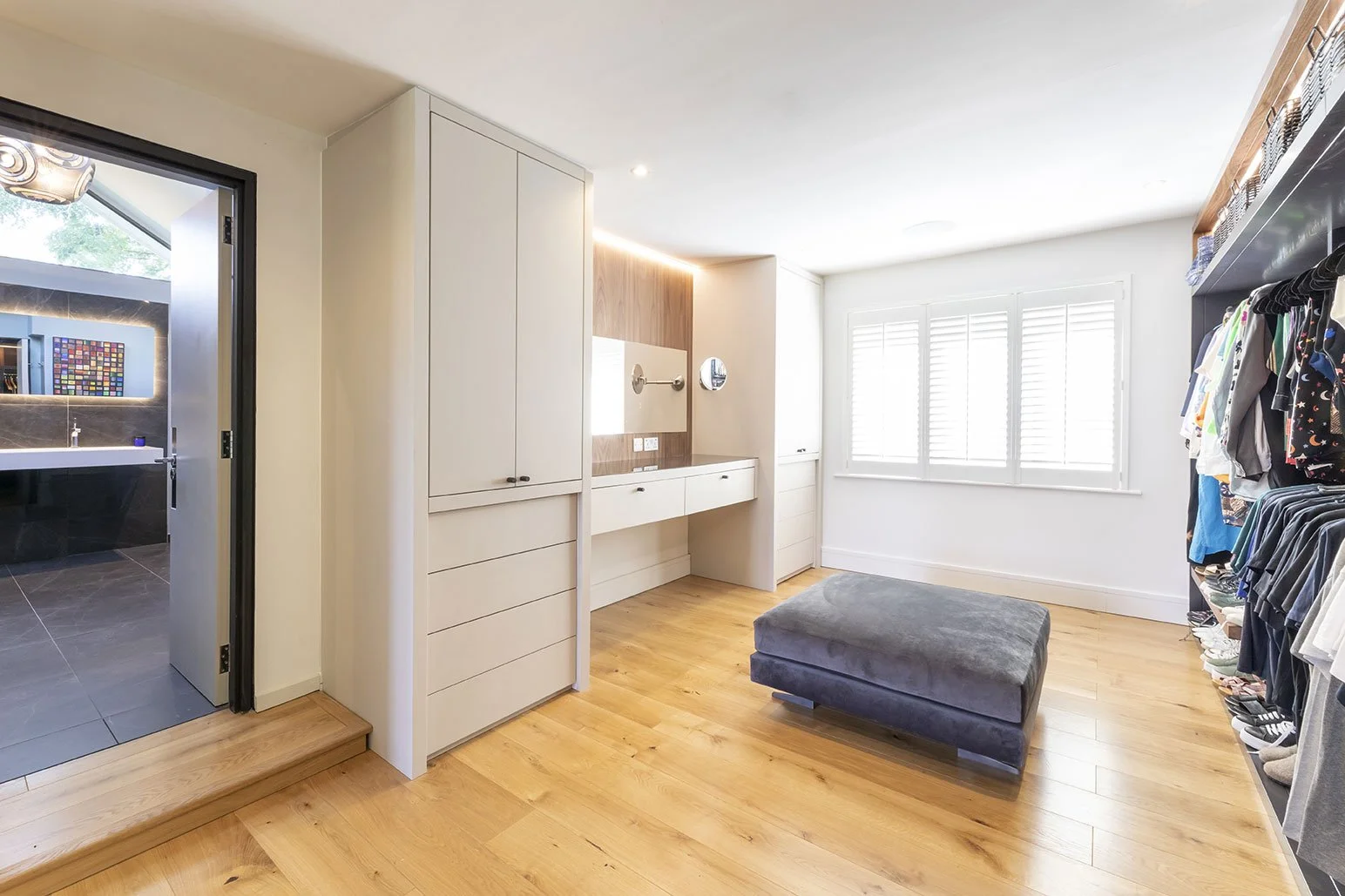 A spacious walk-in closet with wooden flooring, white walls, and large windows with shutters, featuring a gray cushioned ottoman and open shelving with clothes.
