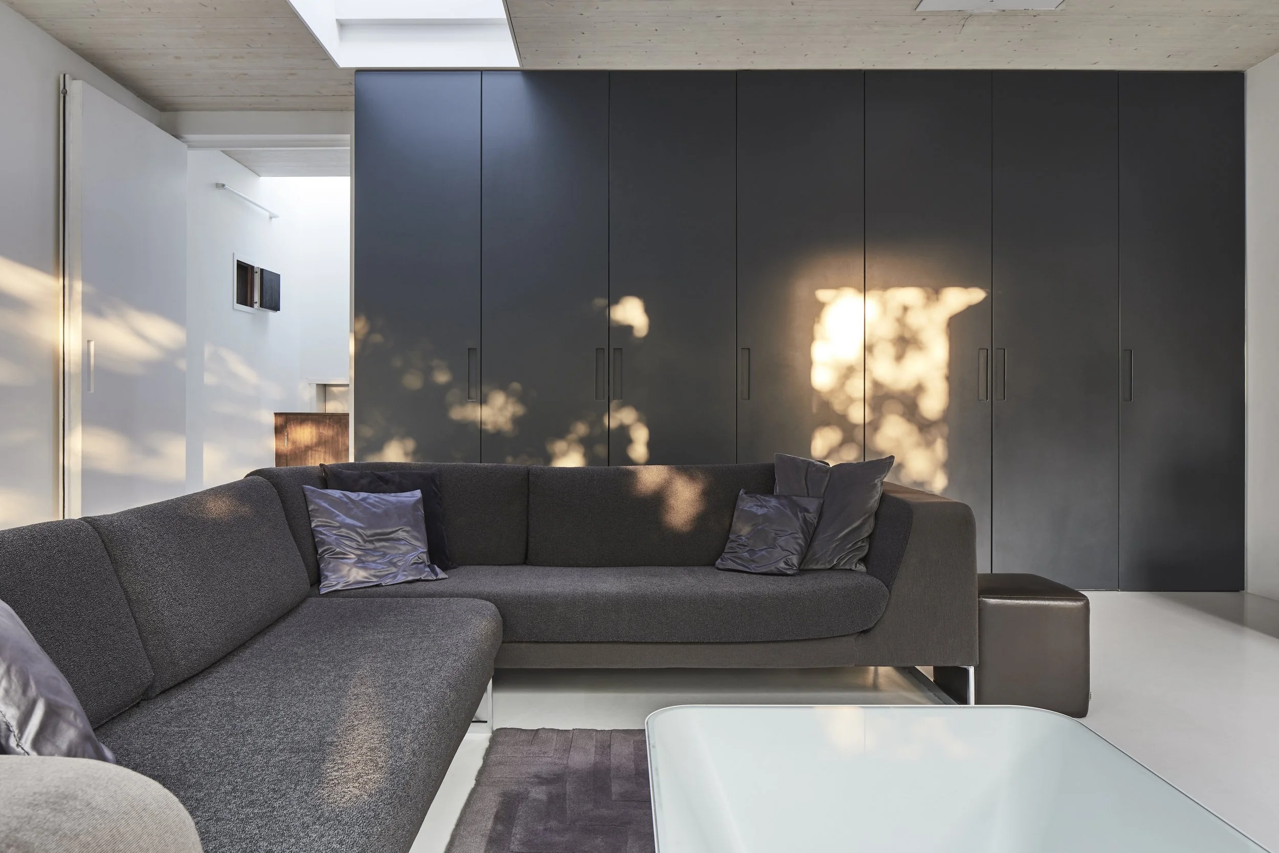 Modern living room with dark gray sectional sofa and black pillows, sunlight casting shadows on a large dark gray wardrobe, white walls and ceiling, and a white coffee table in the foreground.