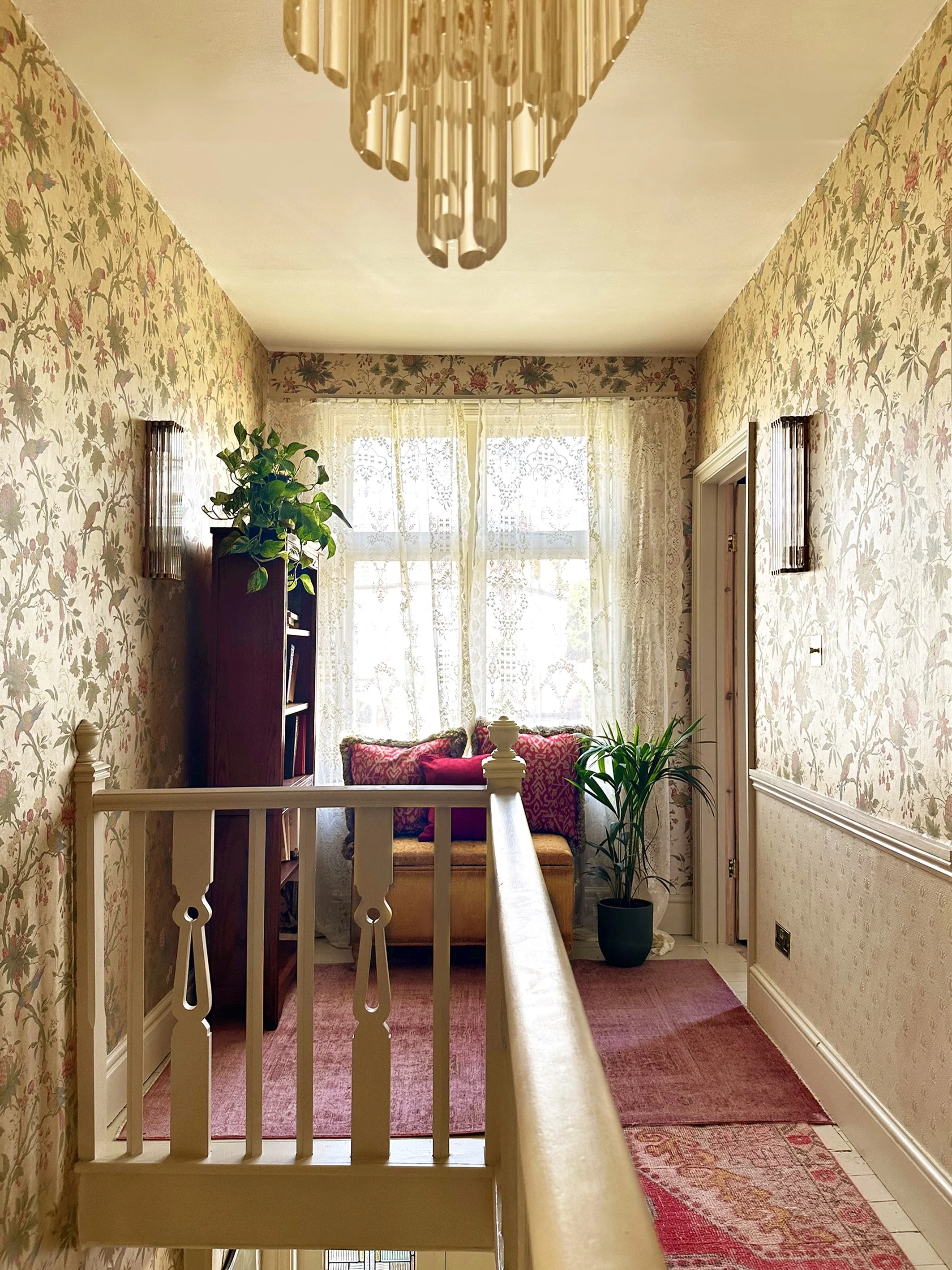 A cozy, vintage-style room with floral wallpaper, lace curtains, a small window seat with cushions, potted plants, and a hanging chandelier.