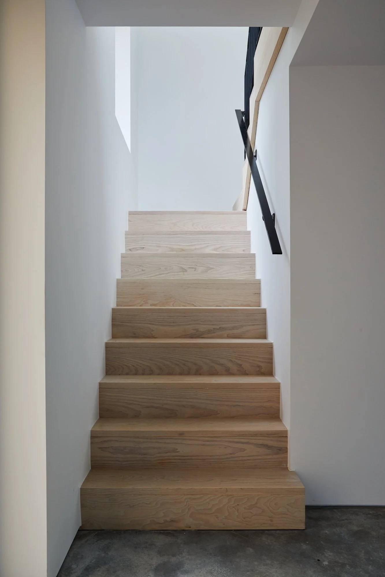 A modern indoor staircase made of light-colored wood with black metal handrails, leading upwards towards a bright, open space with white walls and a skylight above.