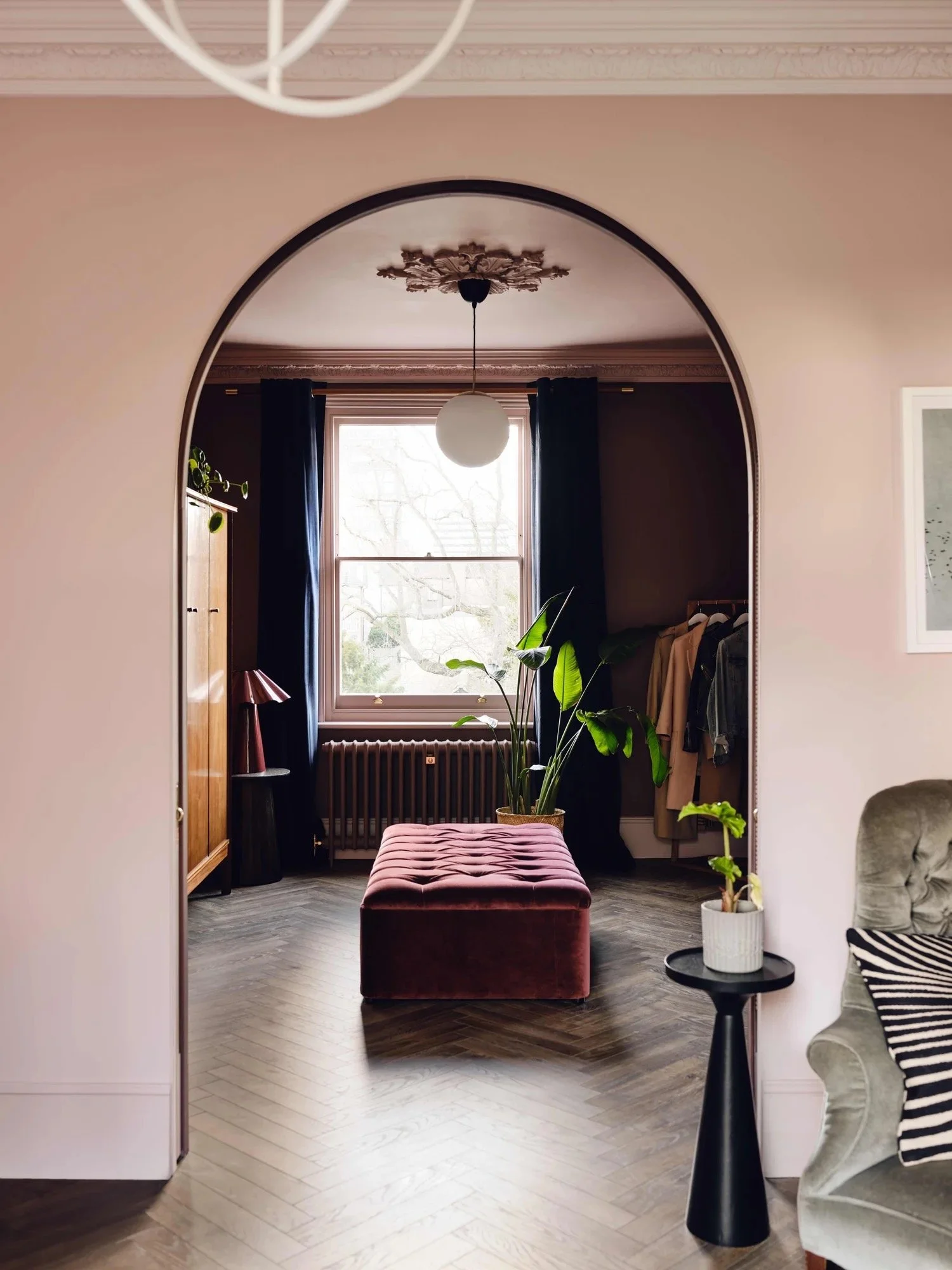 Interior view through an arched doorway showing a room with a window, dark curtains, a large leafy plant, a pink velvet ottoman, a small side table with a potted plant, and a clothing rack with hanging clothes.
