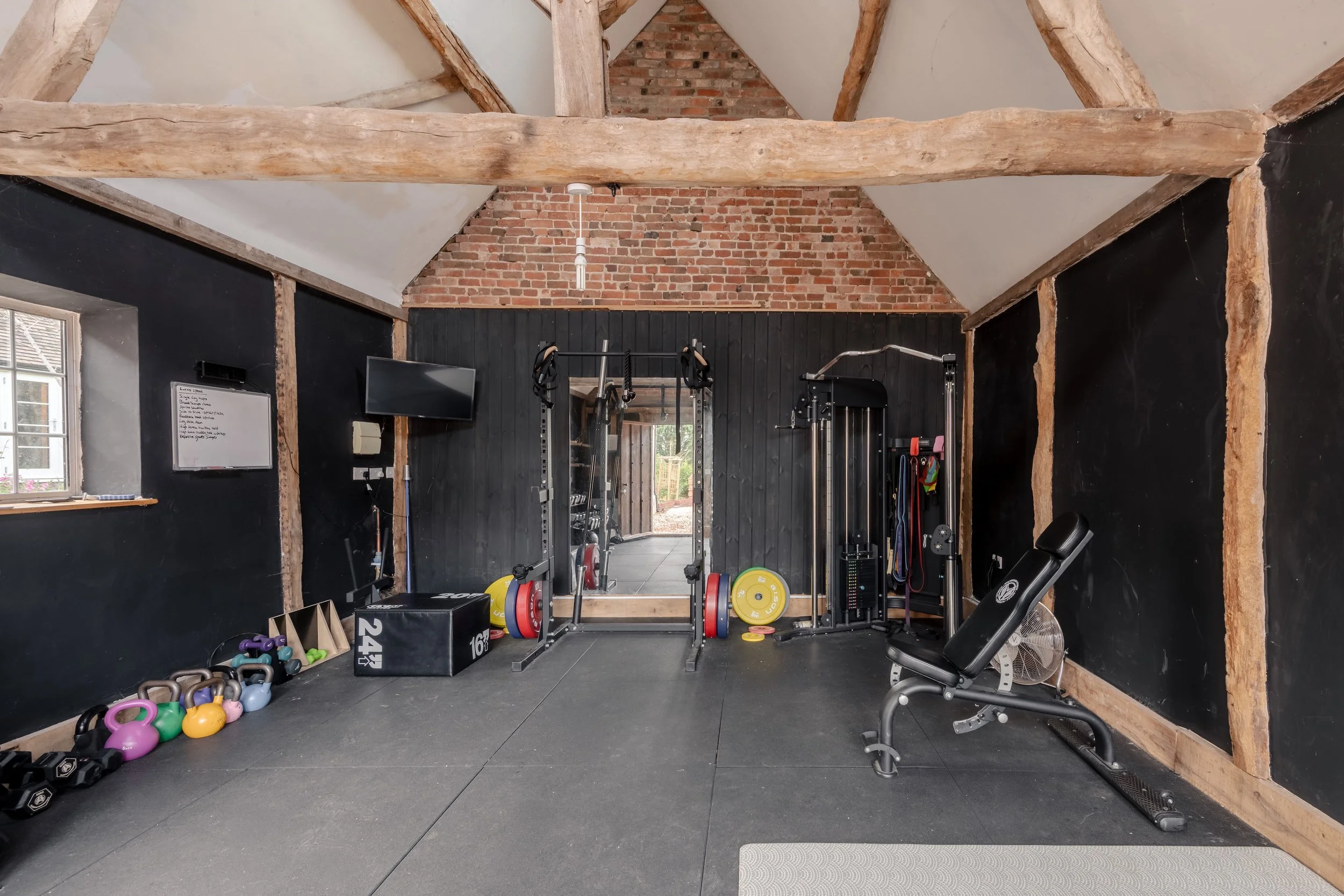 Home gym with black walls, wooden beams, and brick ceiling, equipped with weightlifting equipment, dumbbells, kettlebells, a bench, TV, and mirror.
