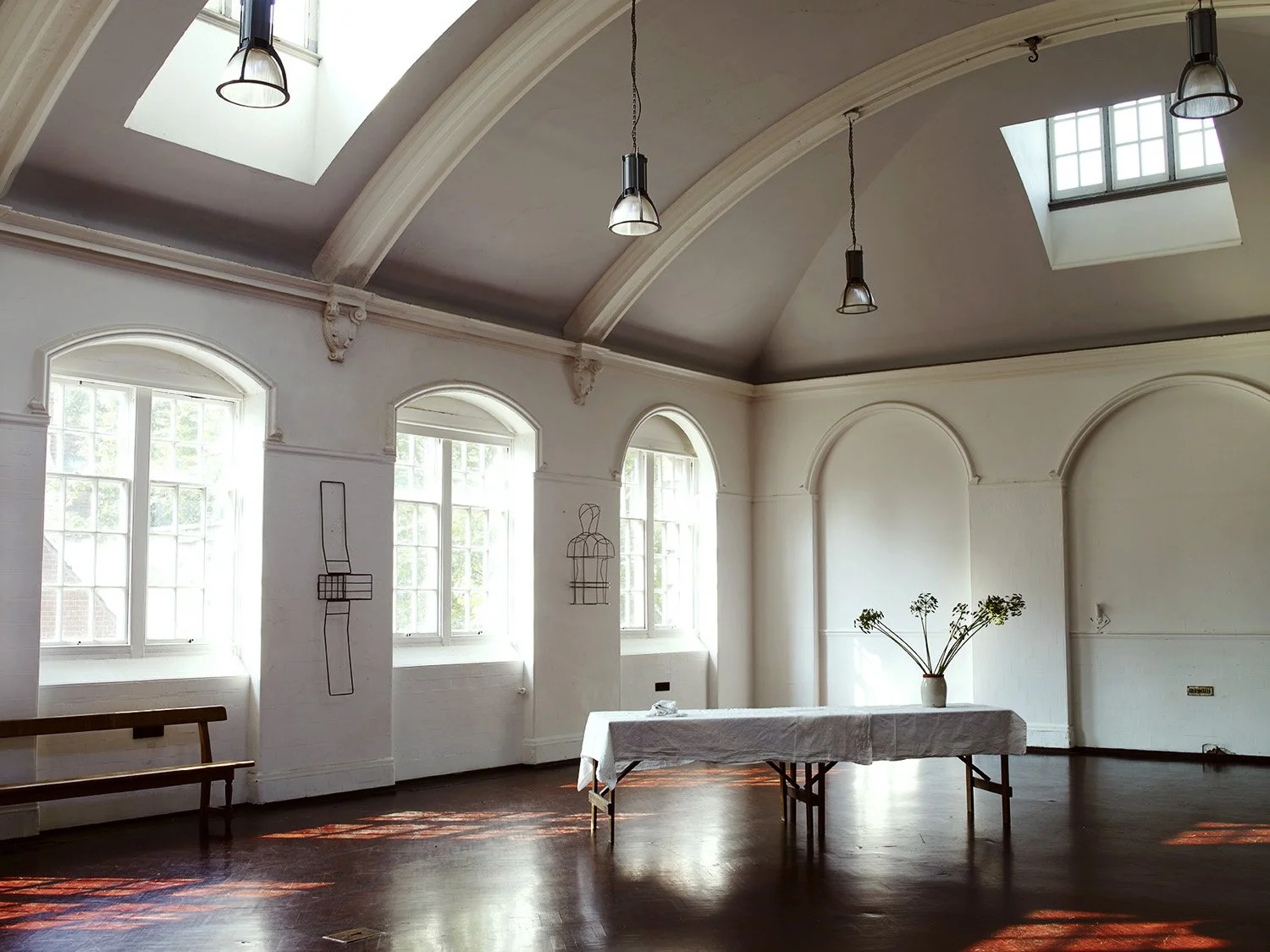 Bright room with high arched ceilings, large windows, and skylights. White walls, dark wood flooring, and minimal furniture including a table with a white cloth and a vase with flowers.