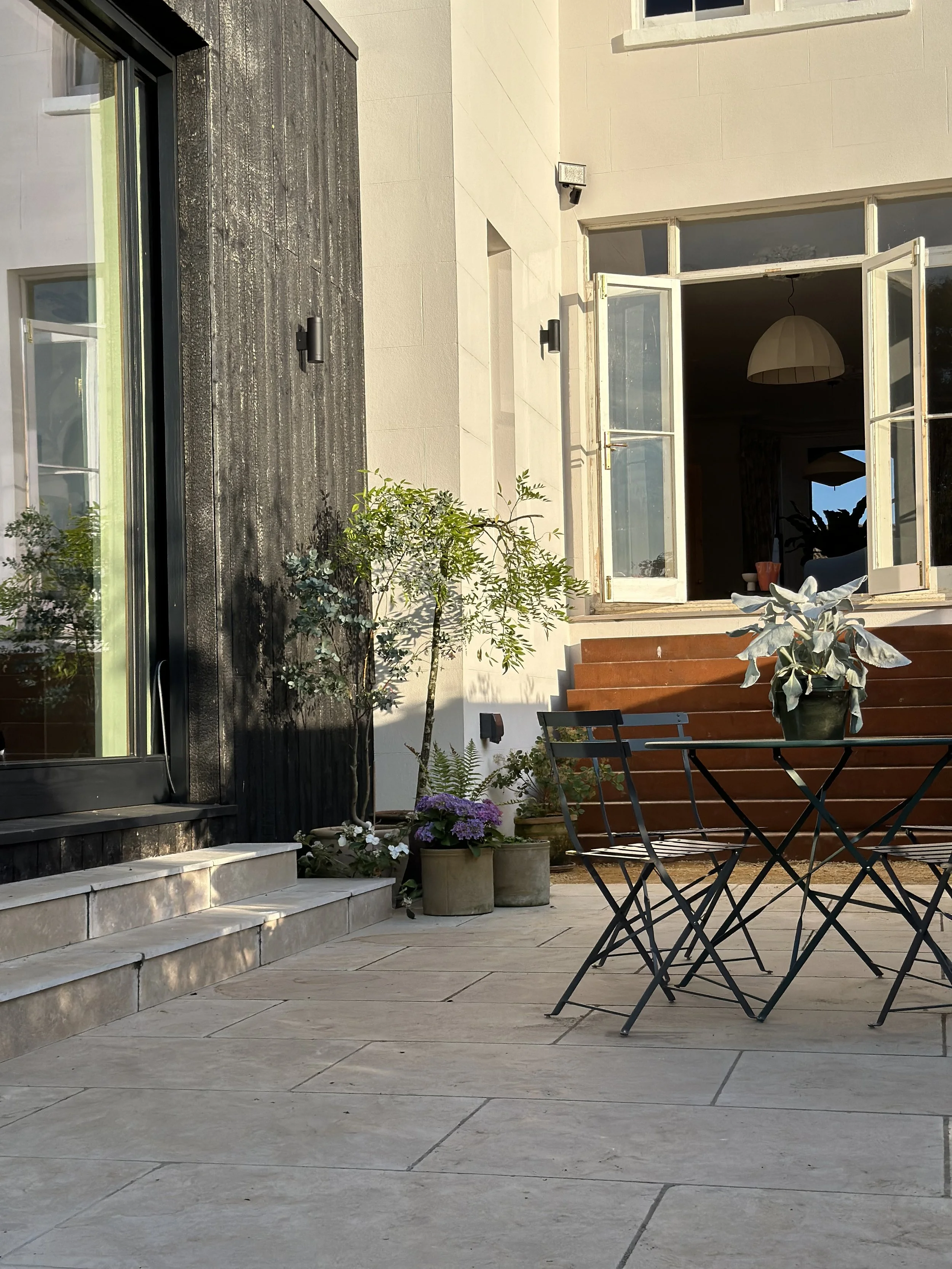 A cozy outdoor patio with a black metal table and chairs, potted plants, and an open door leading into a bright room with a hanging lamp.