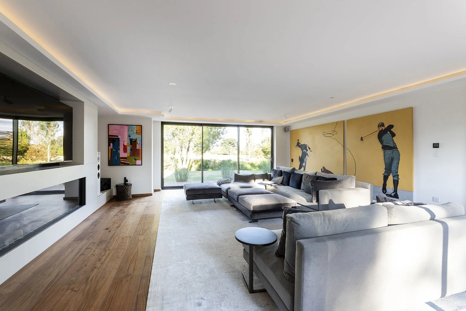 Modern living room with large beige sectional sofa, colorful abstract art on the wall, artwork of a golfer on a yellow background, and sliding glass door leading outside to a garden with trees.