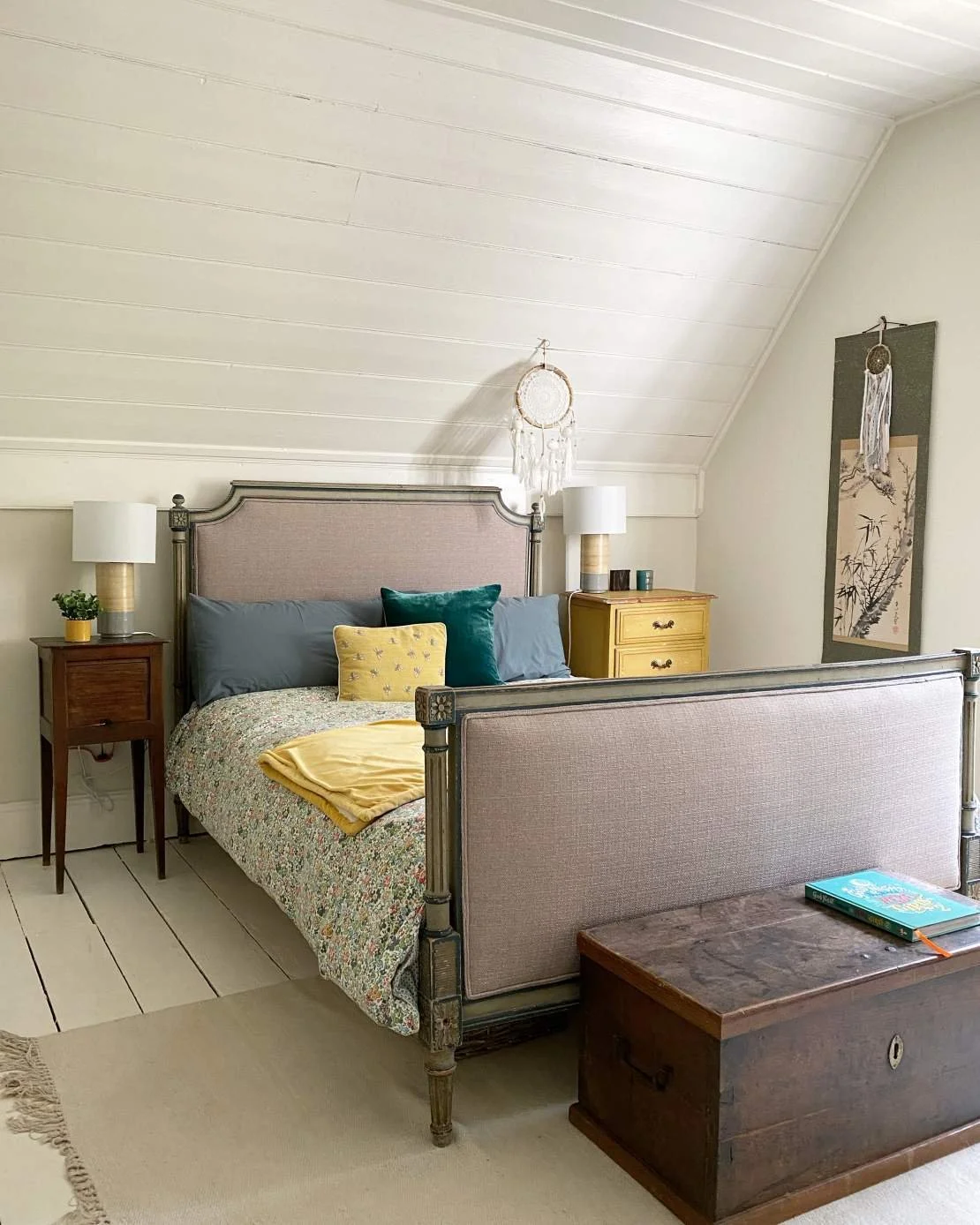 A cozy bedroom with a sloped ceiling, a bed with floral bedding, pillows, and a folded yellow blanket. There are two nightstands with lamps, a chest at the foot of the bed, and wall hangings including a dreamcatcher and Asian-style artwork.