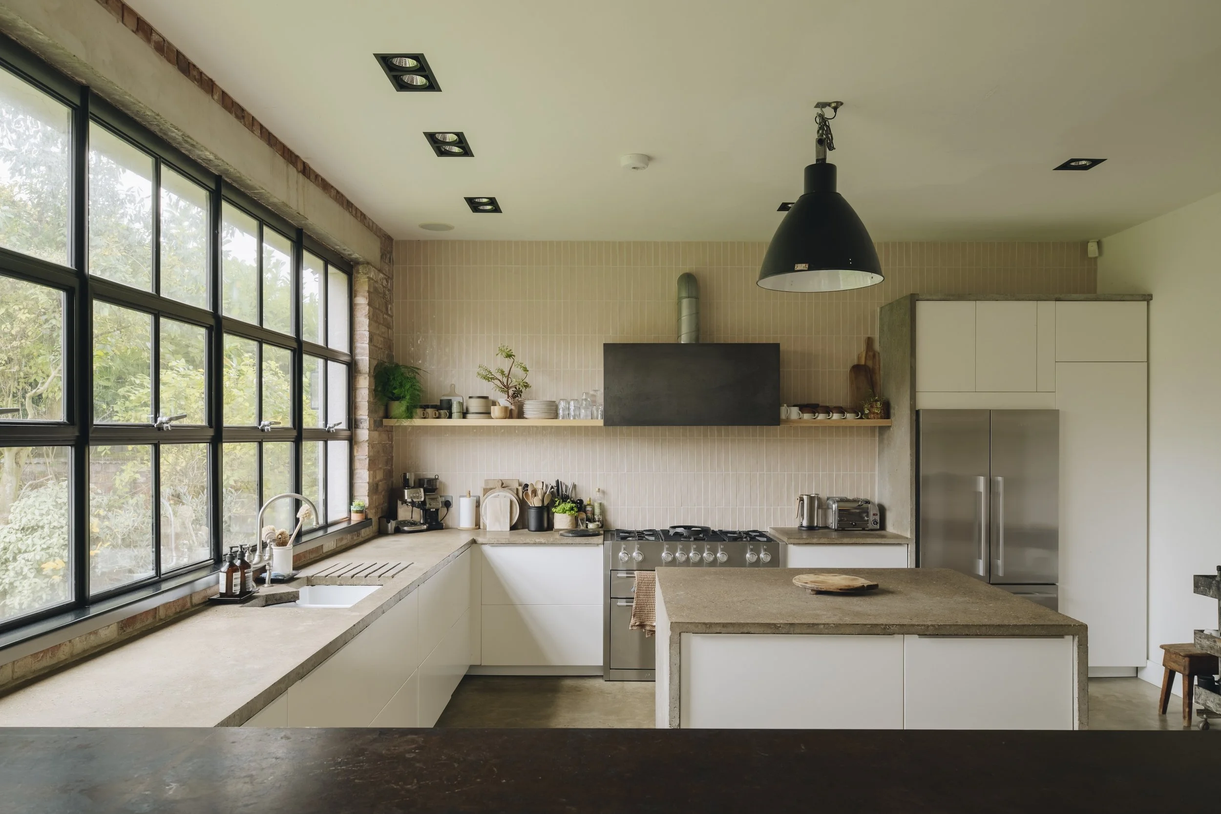 Modern kitchen with large industrial-style black pendant light, beige tiled wall, and big windows with black frames showing greenery outside. Contains white cabinetry, a stainless steel refrigerator, countertop appliances, and open shelves with dishe