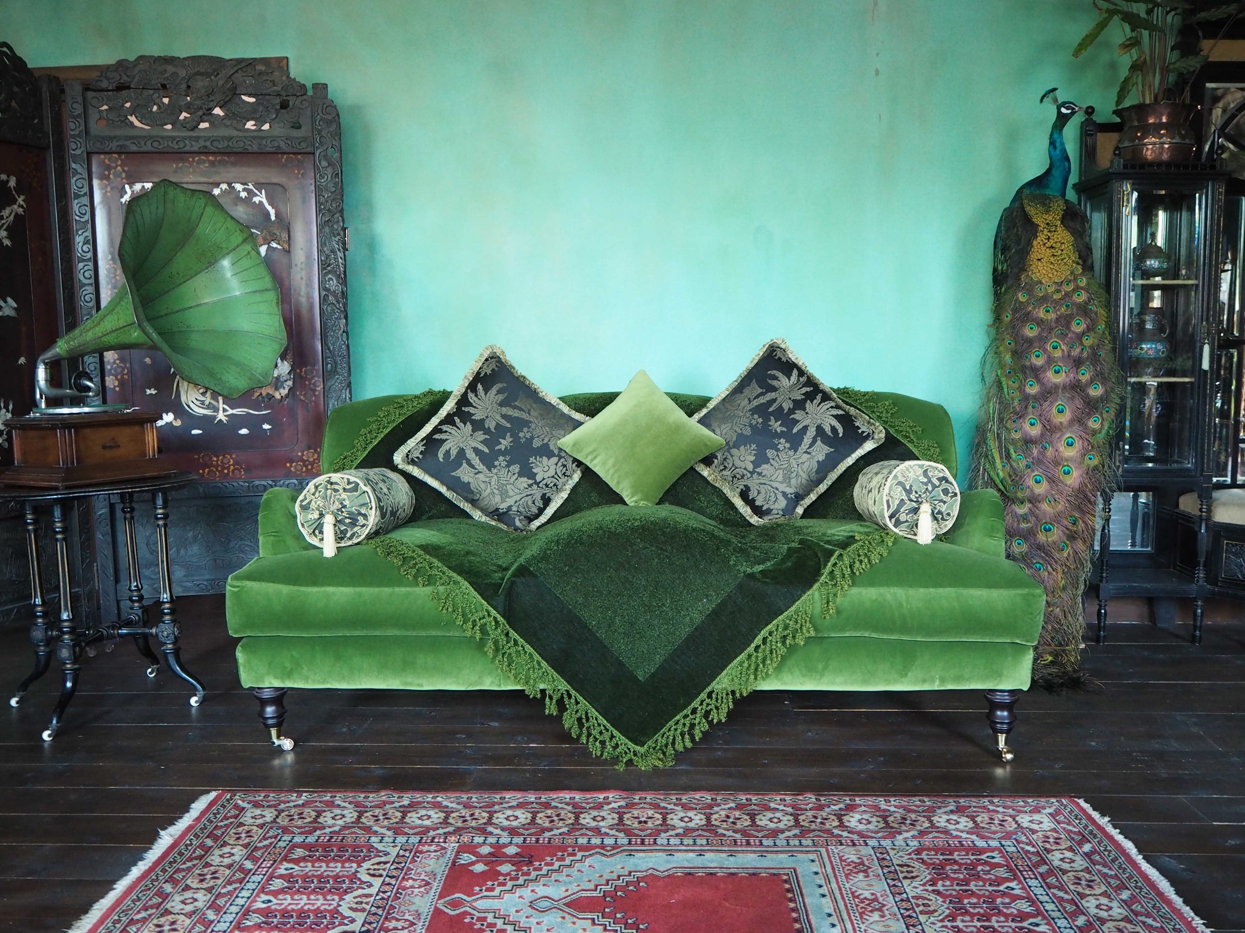 Green velvet sofa with decorative pillows, a large peacock statue, an ornate wooden cabinet, and a Persian rug in a vintage living room.