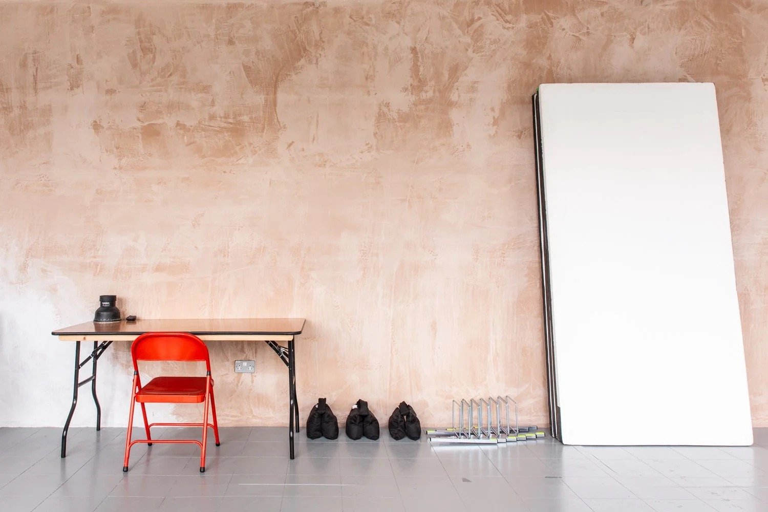 A room with a table, red chair, black bags, a whiteboard, and some floor equipment against a textured peach-colored wall.