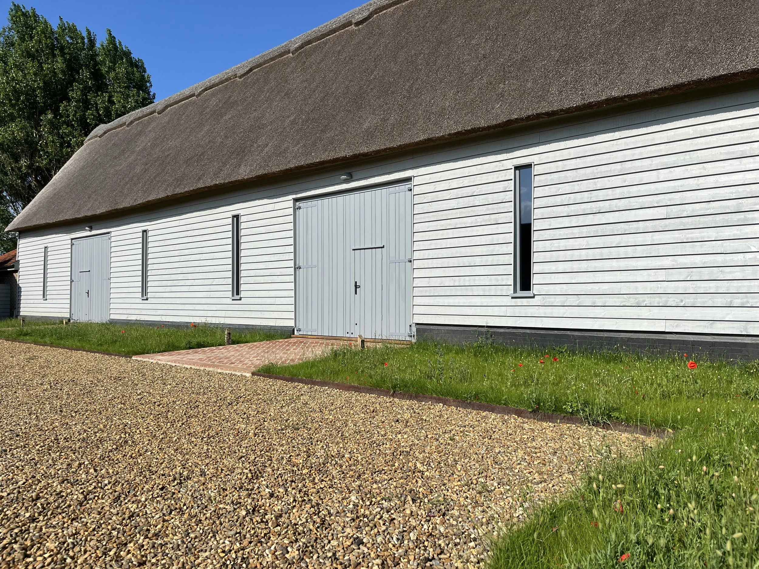 A large white building with a thatched roof, featuring three vertical windows, a central gray wooden door, and a gravel pathway leading up to it. There is green grass with red flowers in front of the building, and a clear blue sky overhead.