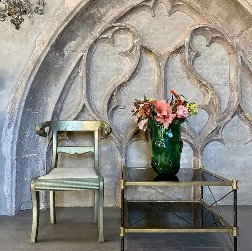 A vintage green upholstered chair and a glass table with gold accents, holding a green vase with pink and peach flowers, set against an ornate stone wall with gothic arches.