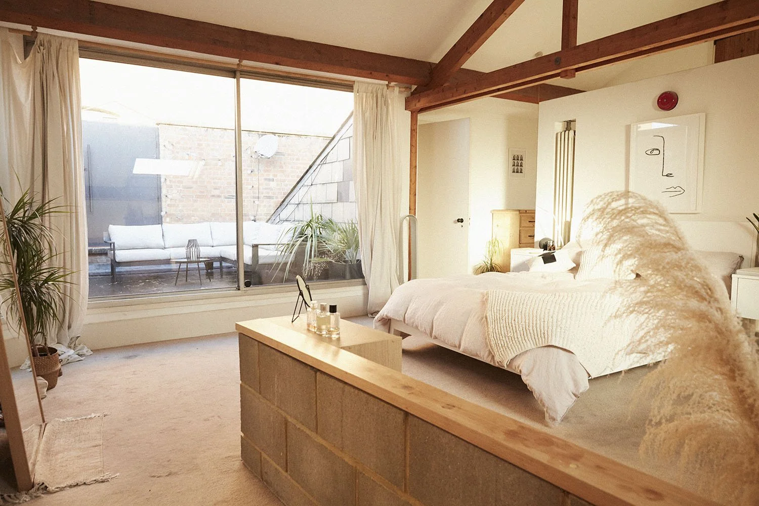 Bright bedroom with large window, white bed with textured pillows, wooden beams, indoor plants, and a patio with outdoor seating.