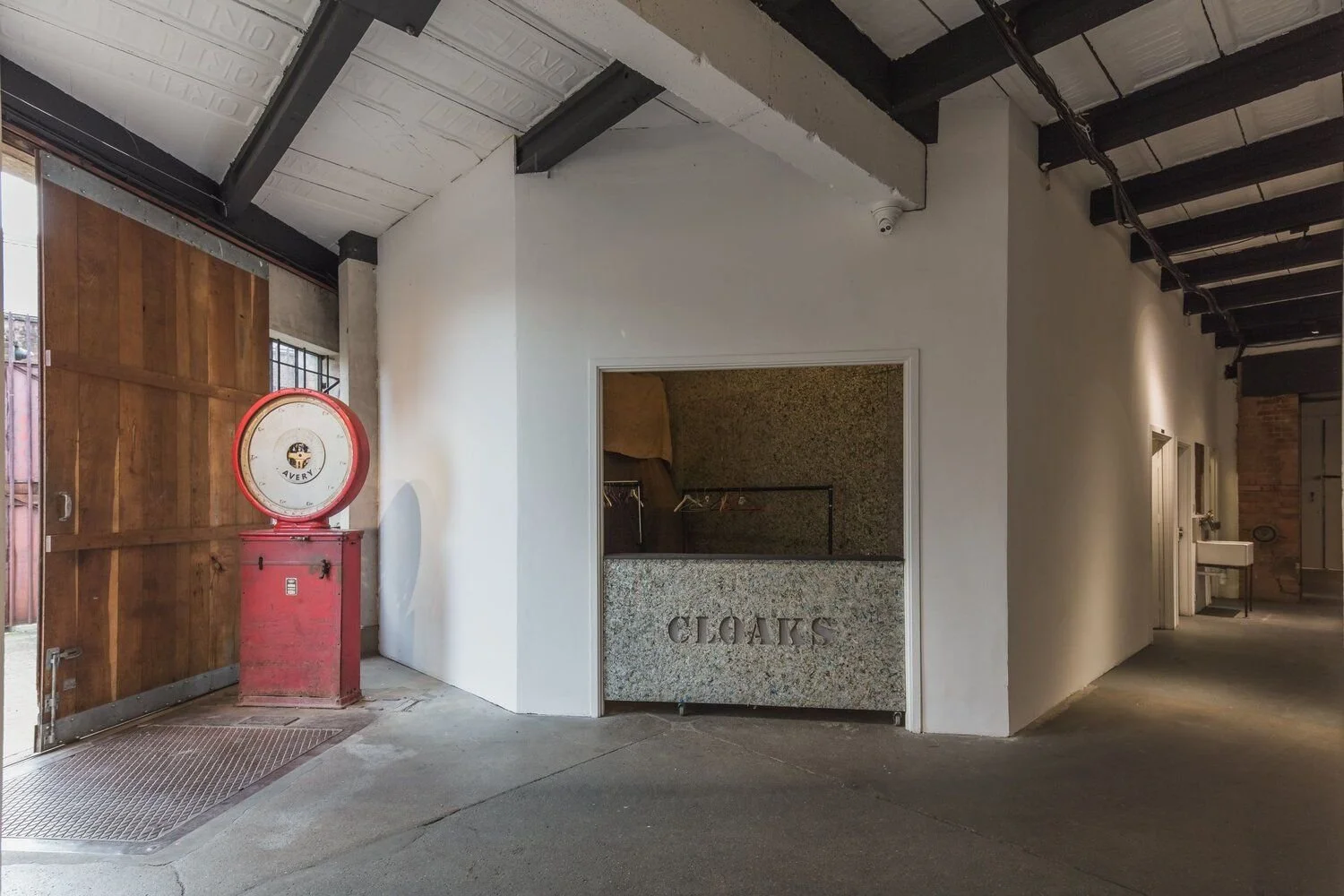 Interior of a building with a vintage red scale near a wooden door and a white wall with a windowed booth labeled 'CLOAKS' in granite.
