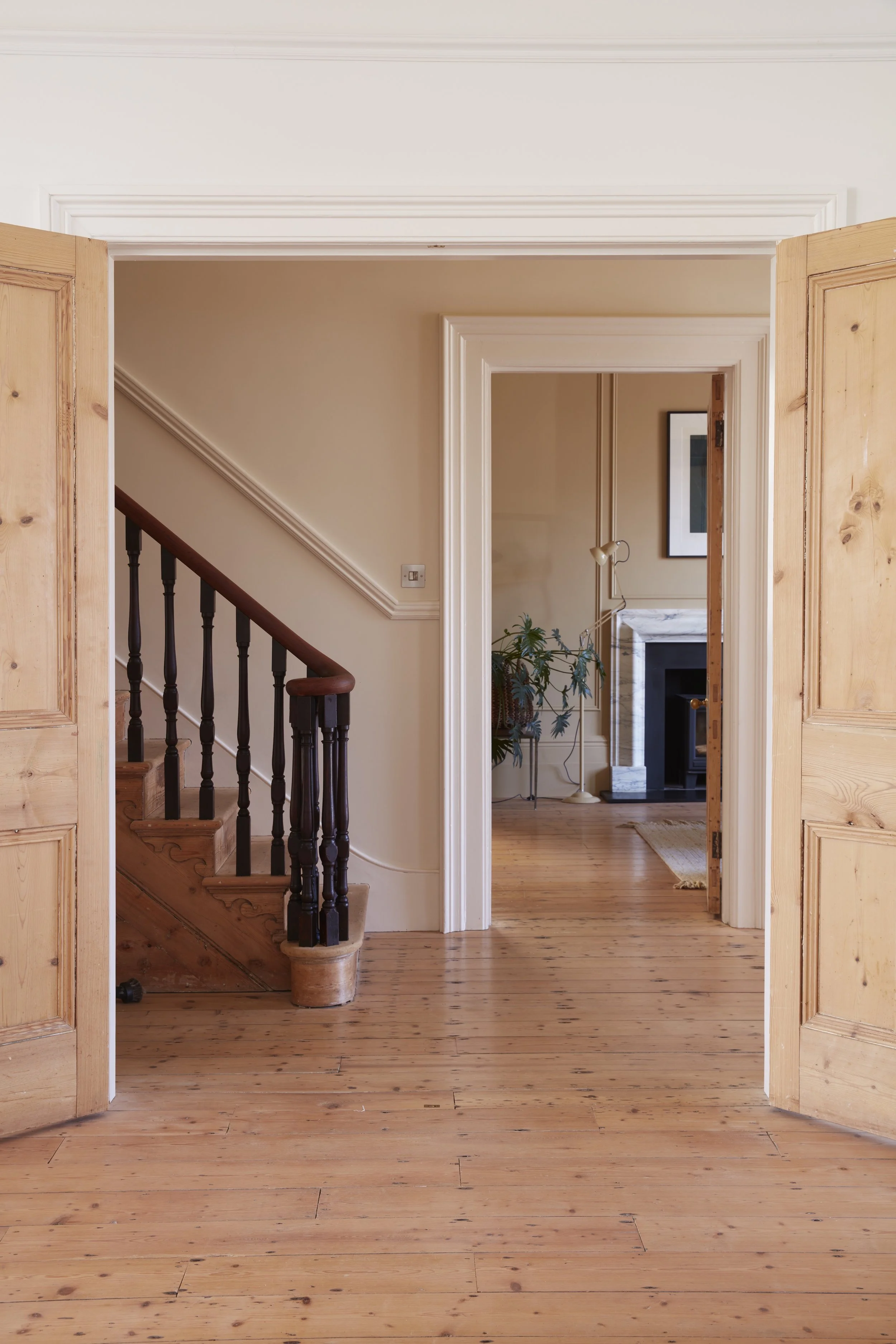 Interior view of a house with wooden floors and open double doors leading to a hallway with stairs, a plant, and a fireplace