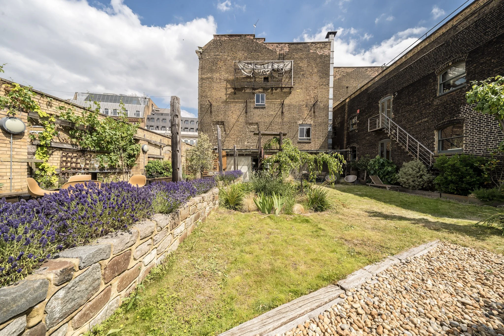 Urban backyard garden with plants, lavender flowers, a stone border, and outdoor furniture against brick buildings and a partially cloudy sky.