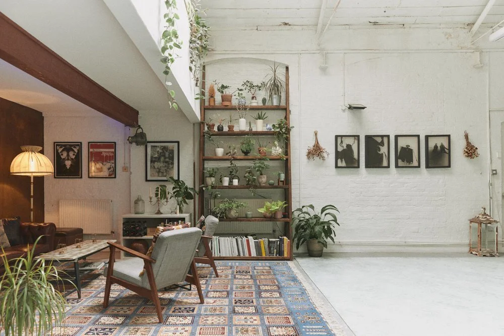A cozy, modern living room with a white brick wall, black and white artwork, potted plants, and a wooden shelving unit with decorative items and books. There is a patterned area rug, a vintage floor lamp, and seating furniture including a beige armch