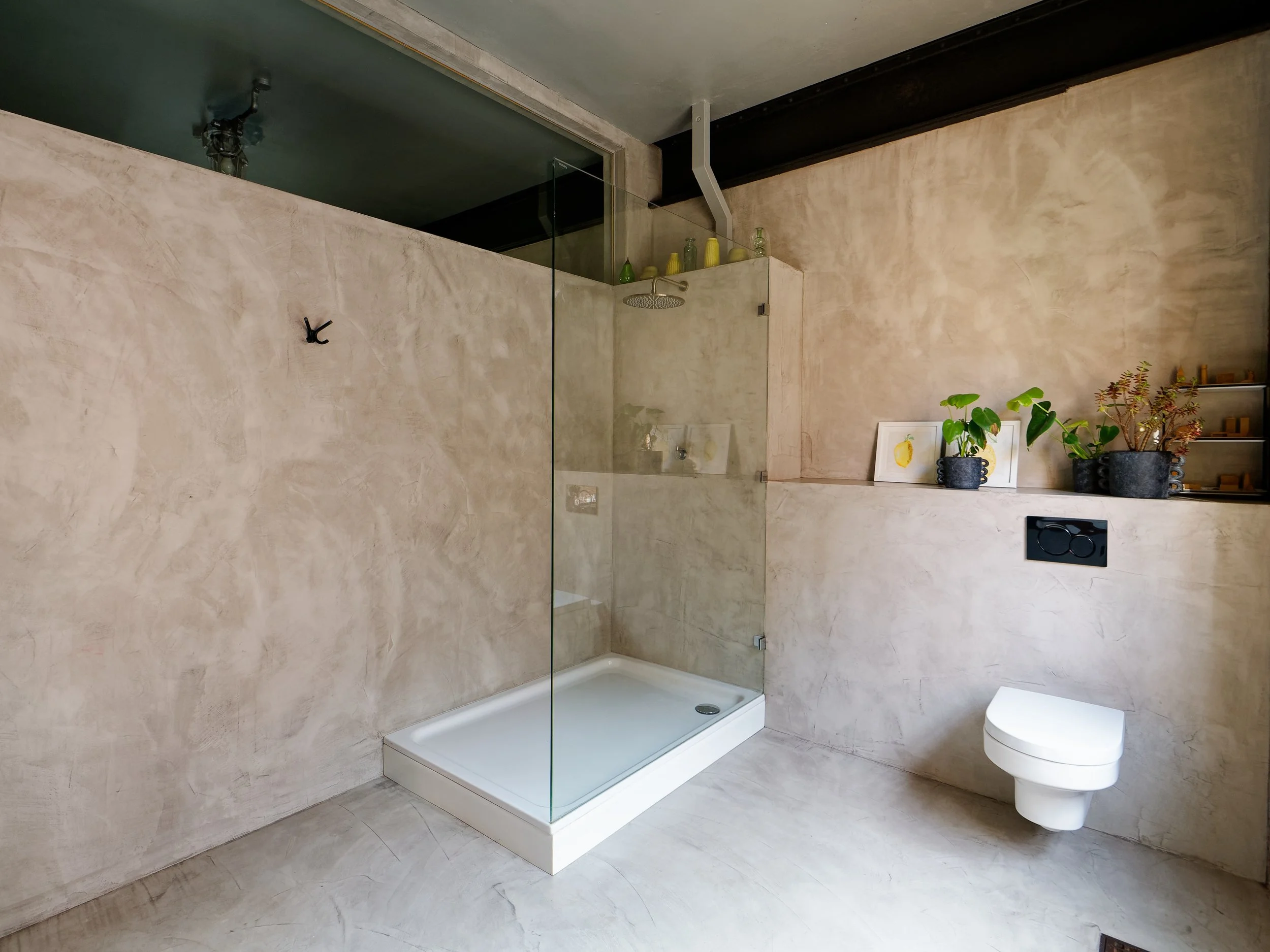 Modern bathroom with a concrete wall finish, a glass shower enclosure with a rain showerhead, a wall-mounted toilet, and a shelf with potted plants and framed artwork.