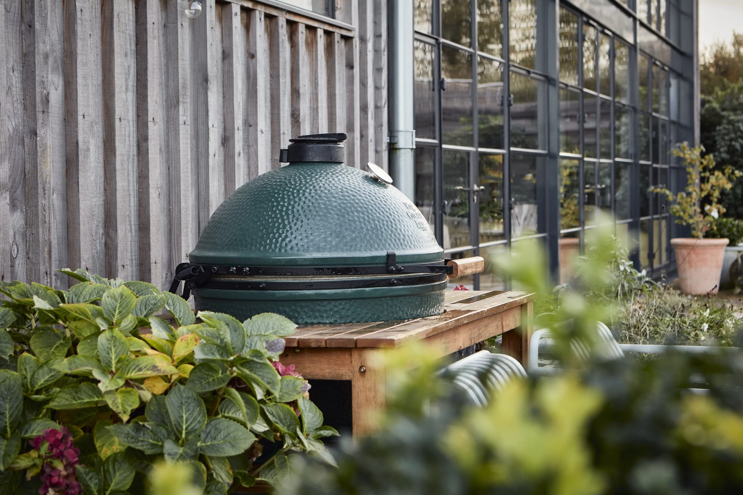 A green, dome-shaped ceramic grill on a wooden table in a garden with plants and flowers, next to a wooden fence and a glass greenhouse.