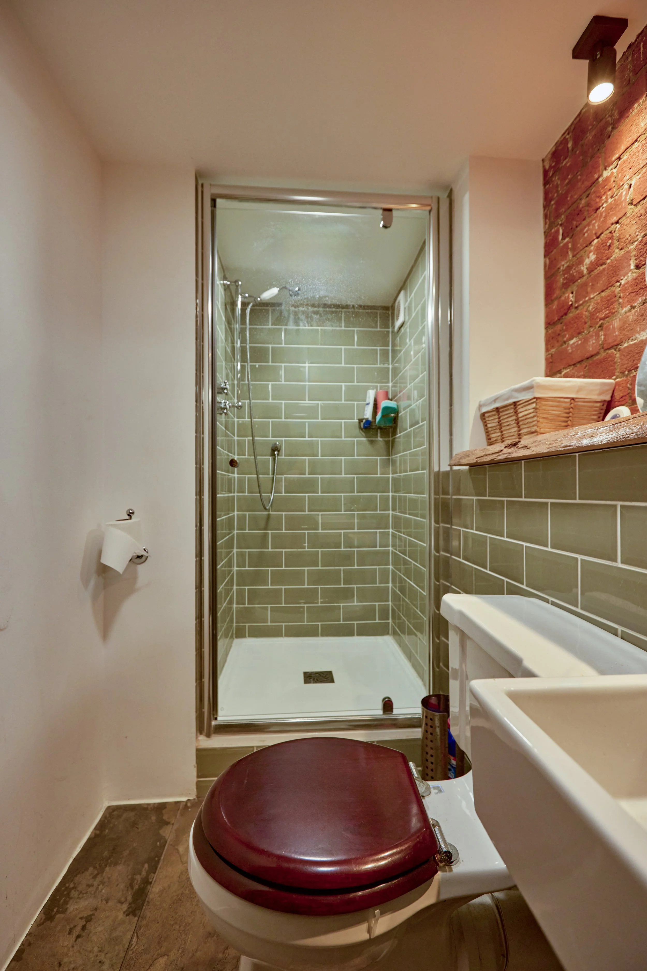 Bathroom with a shower enclosure with green tiles, a burgundy toilet seat, a small sink, and a partially visible wall-mounted mirror. The shower has a shelf with toiletries, and the bathroom has a mixed brick and painted wall.