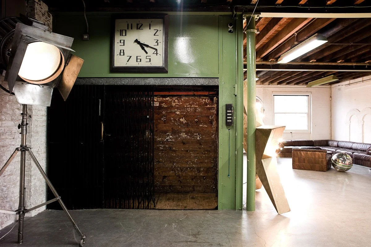 Interior of a studio or lounge space with a large wall clock, studio light on a stand, brick wall, green painted wall, and seating area with a sectional couch, ottoman, and a disco ball.
