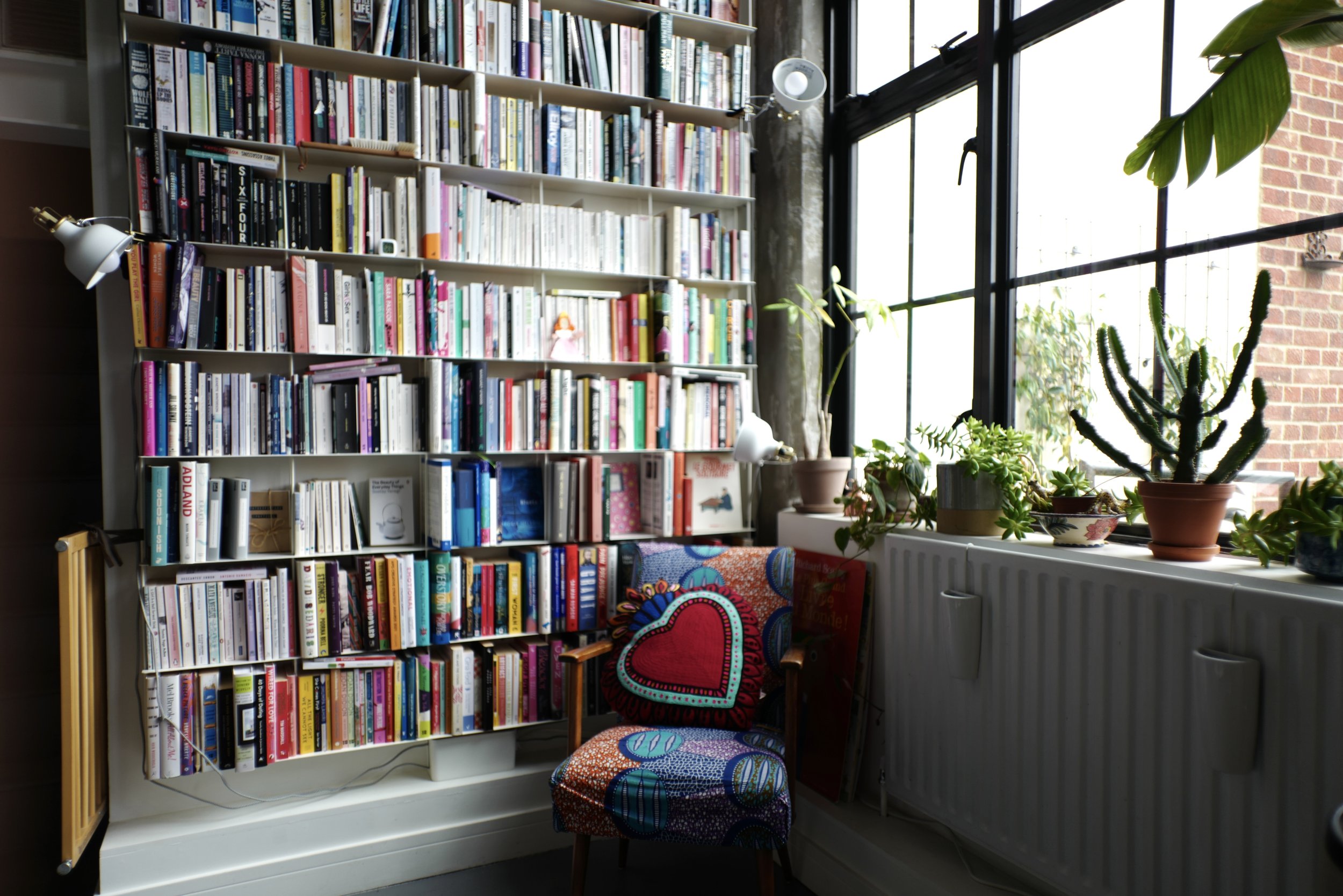 Bright indoor space with a large white bookshelf filled with books, a colorful patterned chair with pillows, potted plants near a large window with black frames, and a radiator beneath the window.