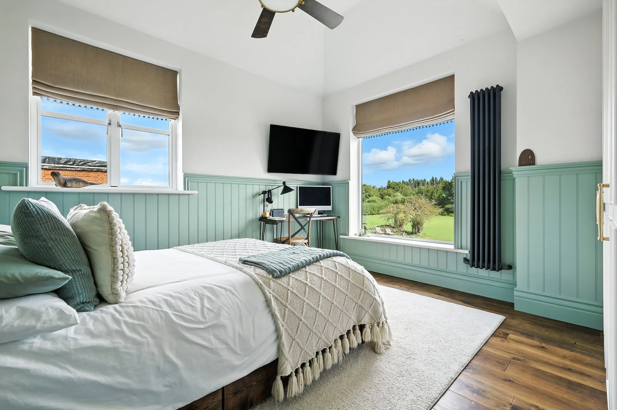 A cozy bedroom with two large windows revealing a sunny outdoor landscape with trees and blue sky. The room features a bed with a white quilt, green pillows, and a decorative blanket. There is a wall-mounted flat-screen TV, a small desk with a comput