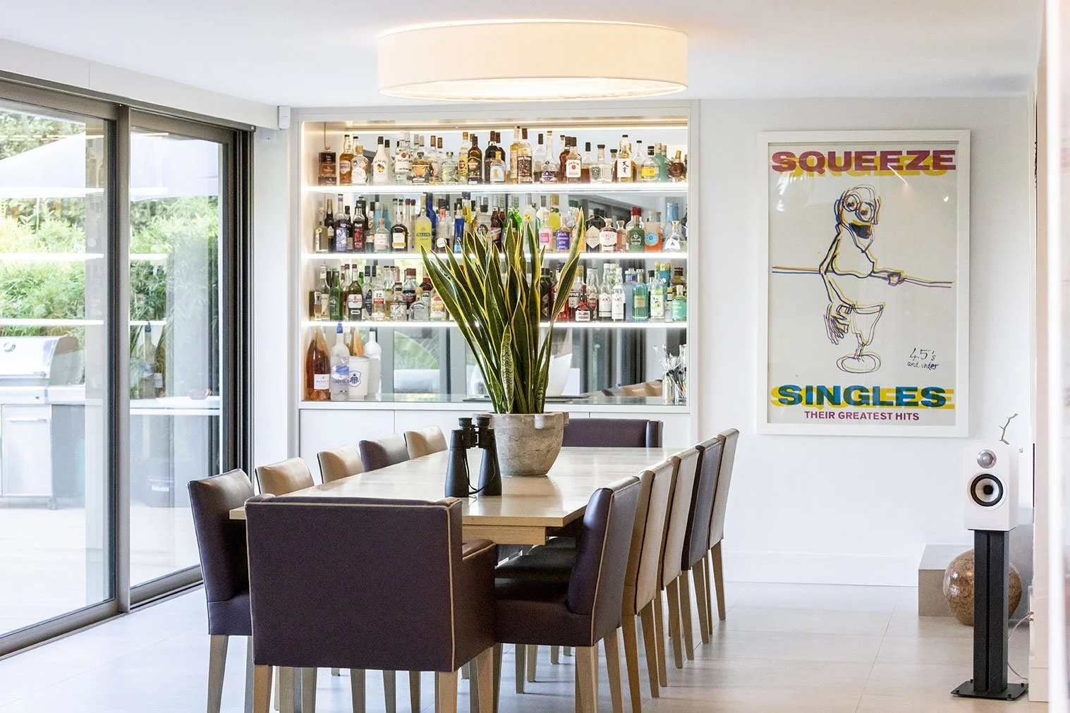 Modern dining room with a wooden table, dark and light chairs, a large potted plant, a bar with various bottles, and a colorful poster on the wall.
