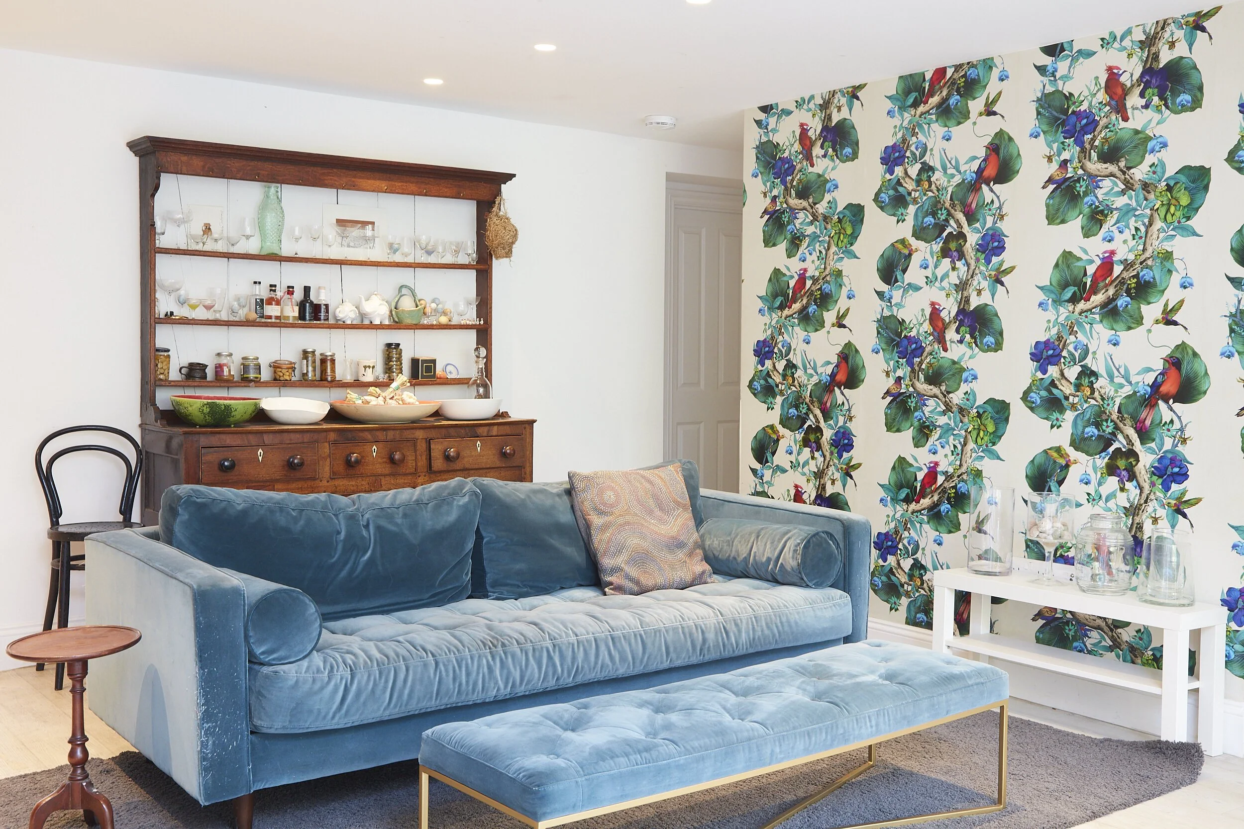 Living room with a blue velvet sofa, a tufted blue ottoman, a floral wallpaper with parrots and flowers, a white side table with glassware, and a wooden hutch displaying dishes and glassware.