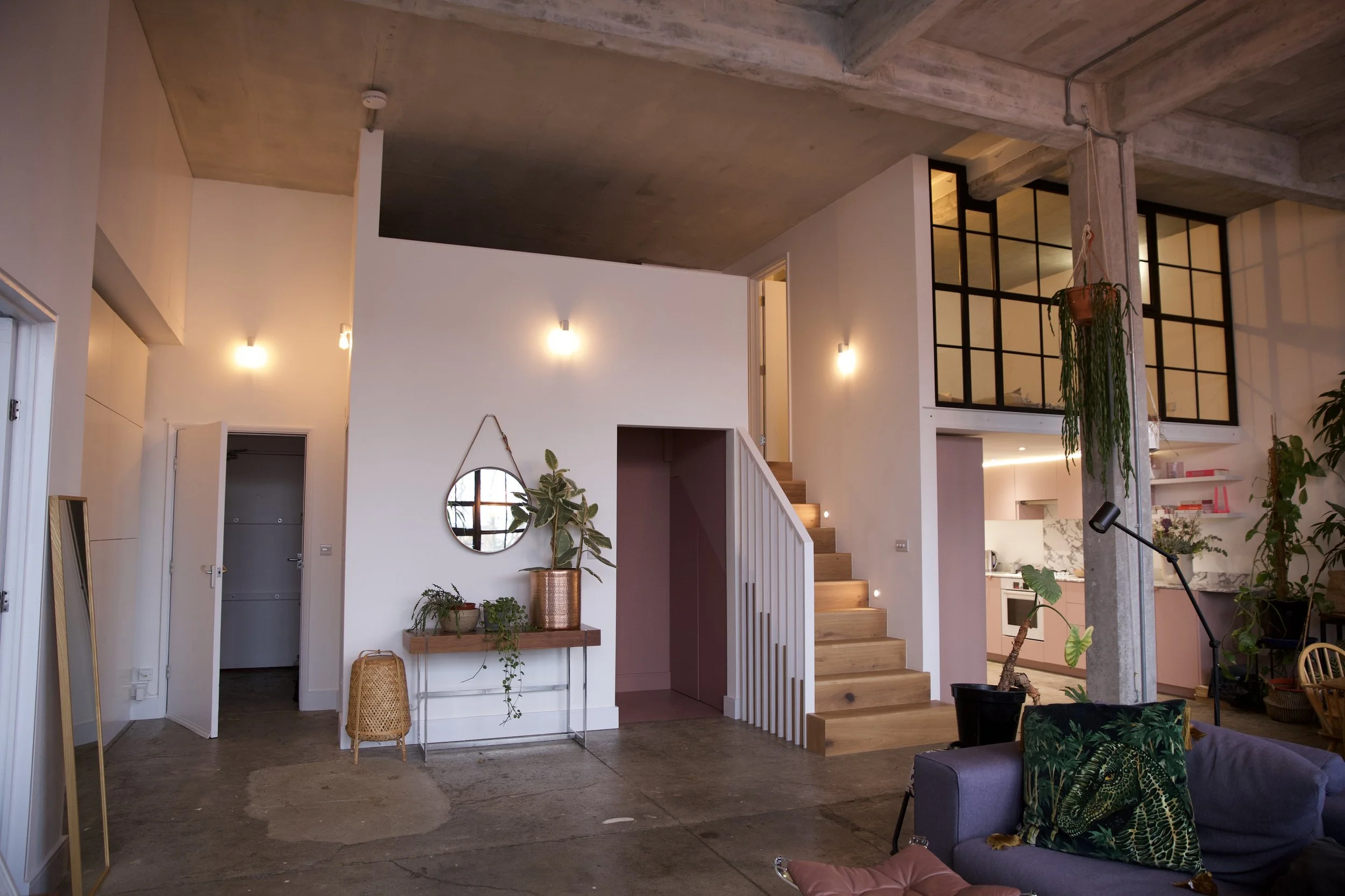 Interior of a modern loft-style apartment with a spiral staircase, exposed concrete ceiling, and plants, including a hanging plant and a potted plant on a console table near a mirror.