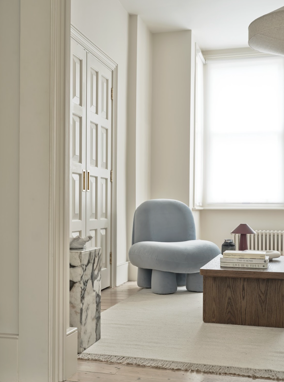 A corner of a living room with a light blue, rounded armchair on a cream rug, a wooden table with stacked books and a small lamp, a window with white curtains, and a white door with brass handles.
