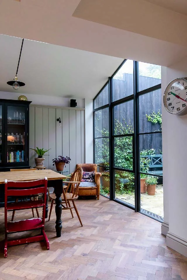 A bright dining area with a wooden table, red, beige, and wooden chairs, a black cabinet with glass doors, and a large glass sliding door leading to a garden with potted plants. A wall clock is visible on the white wall.