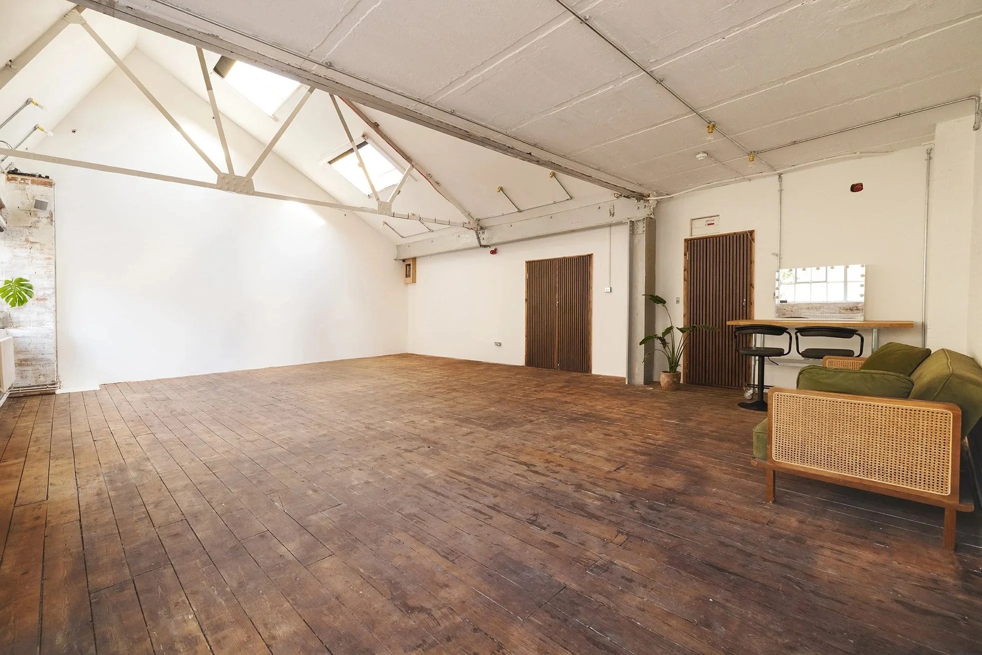 Spacious room with exposed wooden floors, white walls, skylights on a sloped ceiling, and minimal furniture including a green sofa, potted plants, a small bar-height table with stools, and some wooden sliding doors.