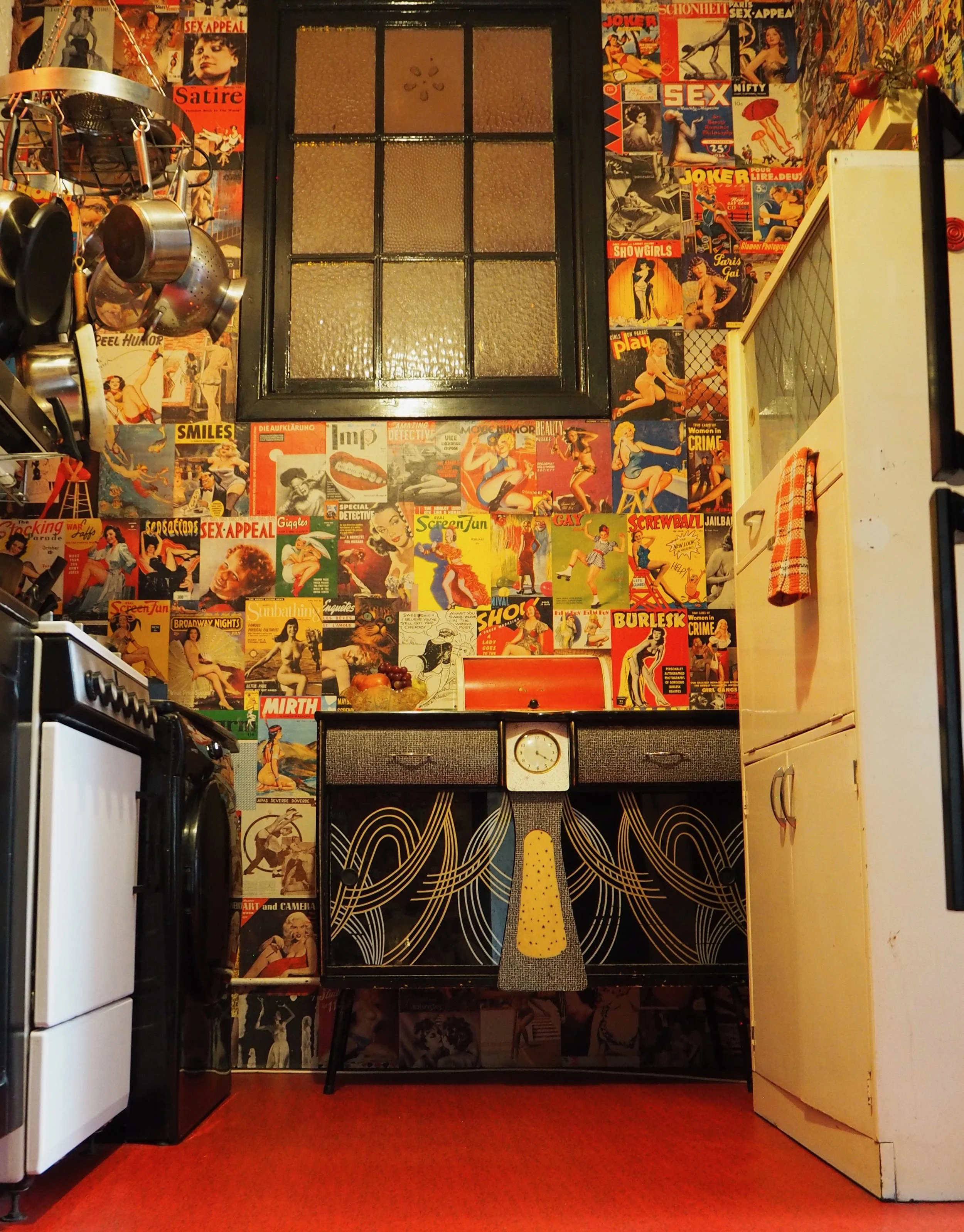 A kitchen decorated with vintage pin-up magazine covers on the wall, a black window, and a small black and gold cabinet with a clock. To the right, a tall white refrigerator with a red and white checkered towel hanging on its handle. To the left, par